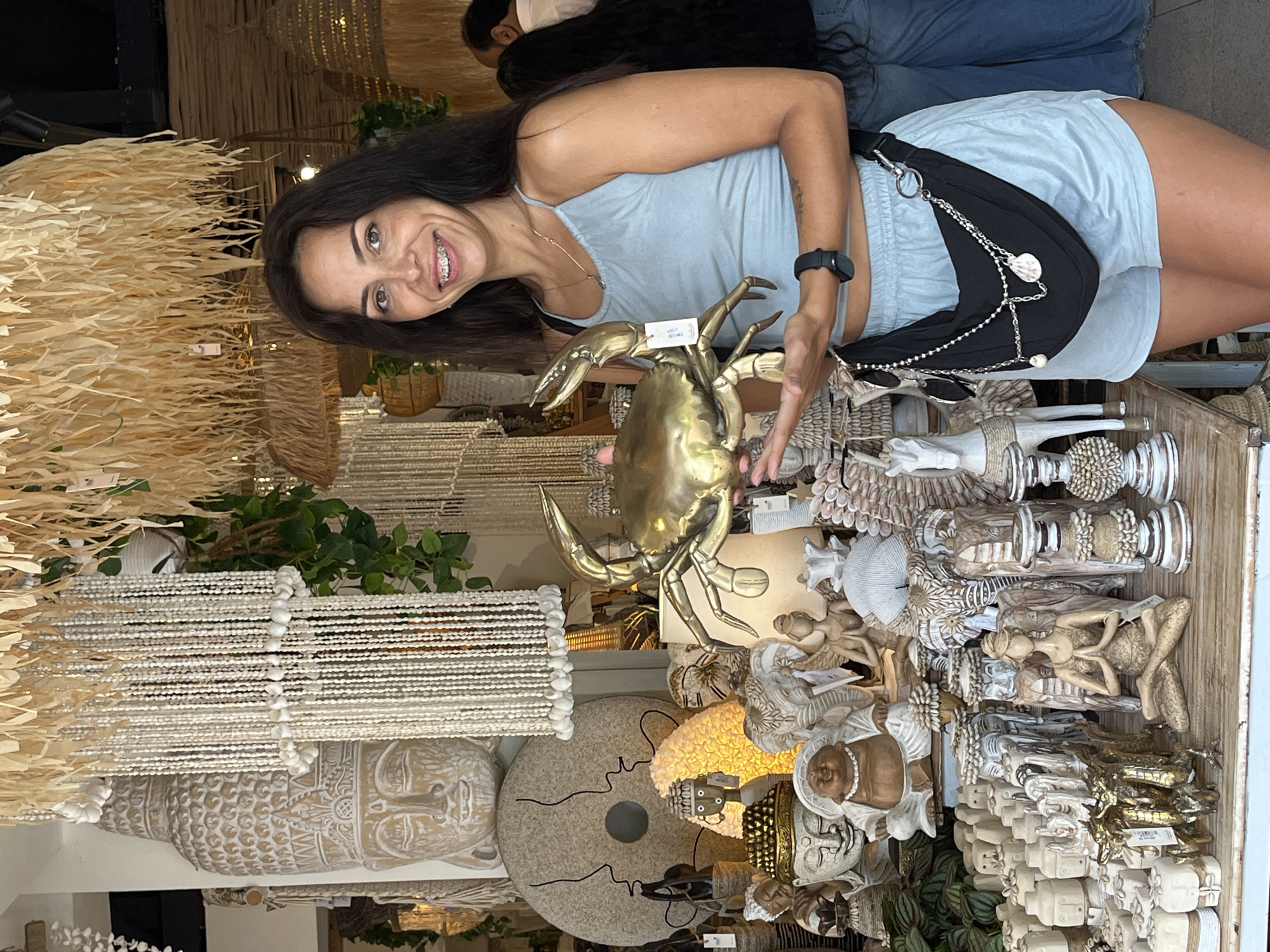 A smiling woman with dark hair and braces, wearing a light blue sleeveless top and matching shorts, is captured indoors in what appears to be a busy market or home decor shop. She holds up a large, polished brass sculpture of a crab with a white tag attached to its claw, looking directly at the camera with a cheerful expression. A black fanny pack or crossbody bag with a chain and shell embellishment is worn across her body, and a black smartwatch is on her left wrist.
The setting is densely filled with a variety of artisanal and decorative items, suggesting a store specializing in cultural or natural-themed home goods, consistent with souvenir shops found in Kuta Selatan, Indonesia. To the left, large textured wall hangings or light fixtures made from natural fibers are visible, alongside a long, hanging decoration crafted from numerous white shells. Below this, a large carved wooden Buddha head is part of the display.
The foreground and midground are packed with an assortment of sculptures and figurines. These include various Buddha statues (some in white, some with gold accents), reclining human figures, elaborate pillars, abstract shapes, and carved wooden or stone objects. One prominent item is a round, grey, textured disc with black lines. Green foliage from potted plants adds a touch of nature to the display. The overall ambiance suggests a vibrant, eclectic marketplace.