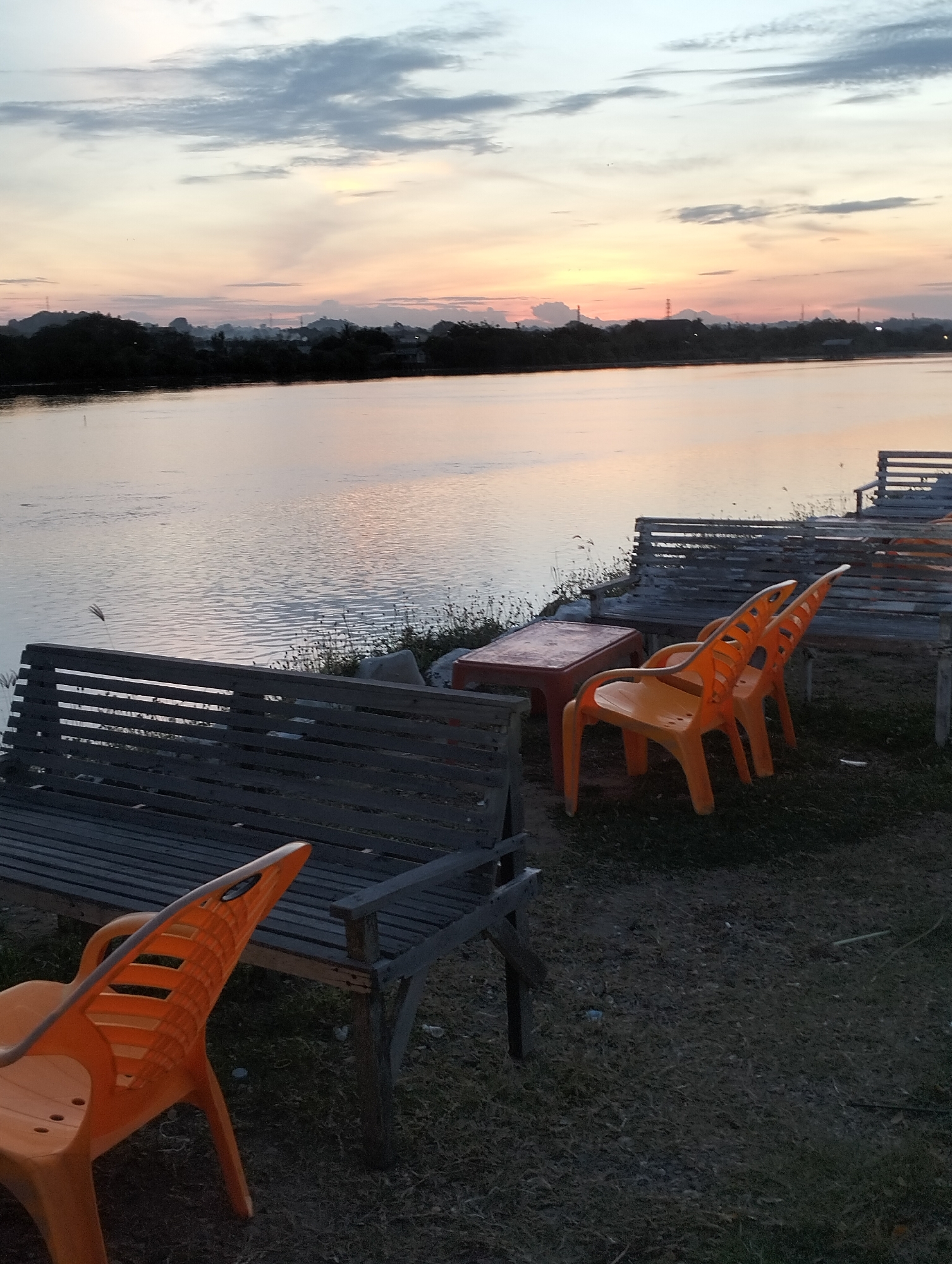 The image depicts a waterfront scene with outdoor seating at sunset, likely in Lhokseumawe, Indonesia, characterized by the following details:
**Setting:** The scene is set by a body of water, possibly a river or bay, with land visible on the opposite shore. The sky is filled with clouds, colored in hues of orange and pink, indicative of either sunrise or sunset.
**Objects:** Wooden benches and orange plastic chairs make up the seating area. A small red table is positioned among the seats.
**Environment:** The immediate foreground is composed of grassy or gravelly ground where the seating is located. The water's surface is rippled, reflecting the colors of the sky.
**People:** There are no people visible in the image.
**Actions/Interactions:** There are no active actions or interactions being captured in this photo. The scene is static.
**Additional Notes:** The image's focus and composition suggest a scenic or travel shot, showcasing the waterfront atmosphere.