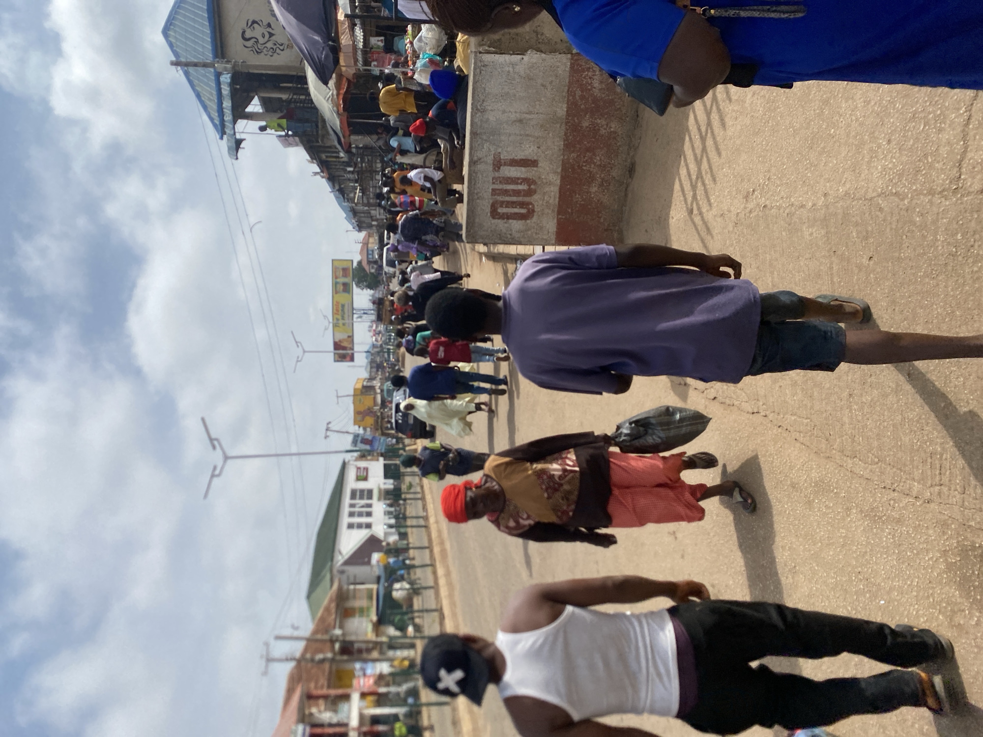 The image depicts a bustling street scene in Bukuru, Nigeria. The perspective is from above, looking down at a busy thoroughfare lined with market stalls and buildings. Several people are visible, walking along the street and in the market area.
In the foreground, three individuals are partially visible. The closest is a person wearing a white tank top and black pants, with a black baseball cap. To their left, a person in a purple t-shirt and denim shorts walks away from the camera. Further down the street, a woman in a patterned dress and a red headscarf is walking, carrying a black bag over her shoulder. Other people, dressed in a variety of casual clothing, are scattered throughout the scene, some of whom appear to be shoppers or vendors.
The setting is an outdoor market or busy street with buildings and stalls on either side of the road. The ground is paved with concrete and asphalt. Overhead, there are power lines and streetlights. The sky is partly cloudy, suggesting it could be daytime, possibly late morning or afternoon, with good visibility.
There is visible text on a concrete barrier in the middle ground, which reads "OUT" in red lettering. There also appears to be a billboard or sign in the background with some text and images. The overall atmosphere seems lively and active, indicative of a typical day in a populated Nigerian town.