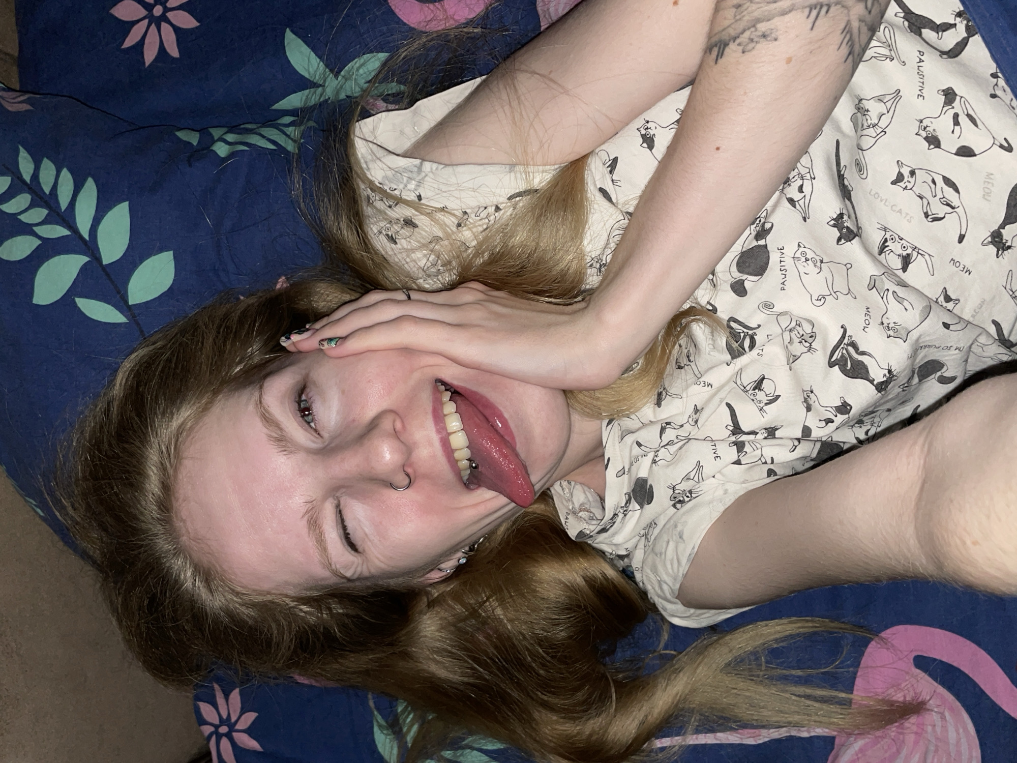 A young woman with light skin and long, light brown hair lies on a blue and pink patterned bedsheet. She is wearing a white short-sleeved shirt patterned with black and white cats. She is making a face, winking with one eye and sticking out her tongue, and has her hand on her face. She has a nose ring and a tongue piercing. The location, Izhevsk, Russia, cannot be determined from the image.