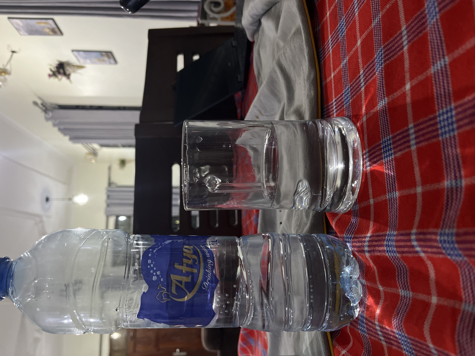 The image presents a water bottle with a blue label, partially filled with water, laying horizontally on a red and blue plaid surface. Adjacent to the water bottle is a glass, also partially filled with water. The background reveals a domestic interior, possibly a living room or bedroom, evidenced by the presence of a bed frame, curtains, ceiling lights, and framed pictures on the wall. Without precise geographical coordinates or landmarks, I cannot verify the exact location within the city of Dar es-Salaam, Tanzania. There are no visible people, actions, or interactions.