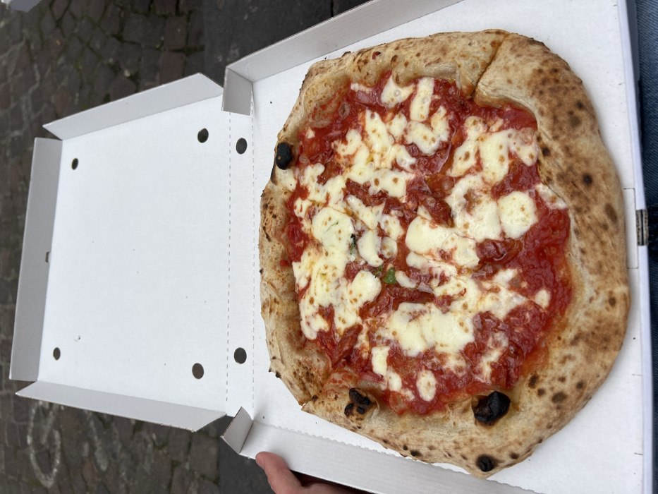 The image shows a Neapolitan pizza in a white cardboard pizza box. The pizza is topped with tomato sauce, mozzarella cheese, and some green peppers. The pizza is in a pizza box sitting on the lap of a person wearing blue jeans. The image was taken in Naples, Italy, at the coordinates 40.842 latitude and 14.251 longitude. The image was likely taken by someone eating the pizza.