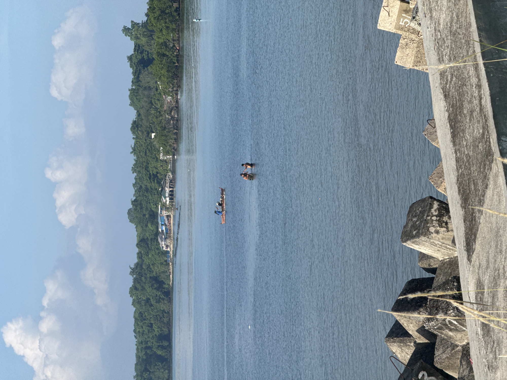 The image depicts a coastal scene in Davao City, Philippines, likely in the area of Sta. Ana pier or a nearby waterfront. The foreground is dominated by large, weathered concrete breakwaters, some marked with numbers like "51". Beyond the breakwaters lies a calm, blue body of water.
In the middle ground, two individuals are wading in the water, appearing to interact with each other. Further out, a small, orange boat with at least two people on board is being rowed. The boat appears to be a traditional outrigger canoe.
The background features a densely forested coastline with lush green trees. Several houses and buildings are visible along the shoreline, suggesting a populated area. The sky is a clear blue with scattered white clouds. The overall impression is of a serene day in a coastal community.