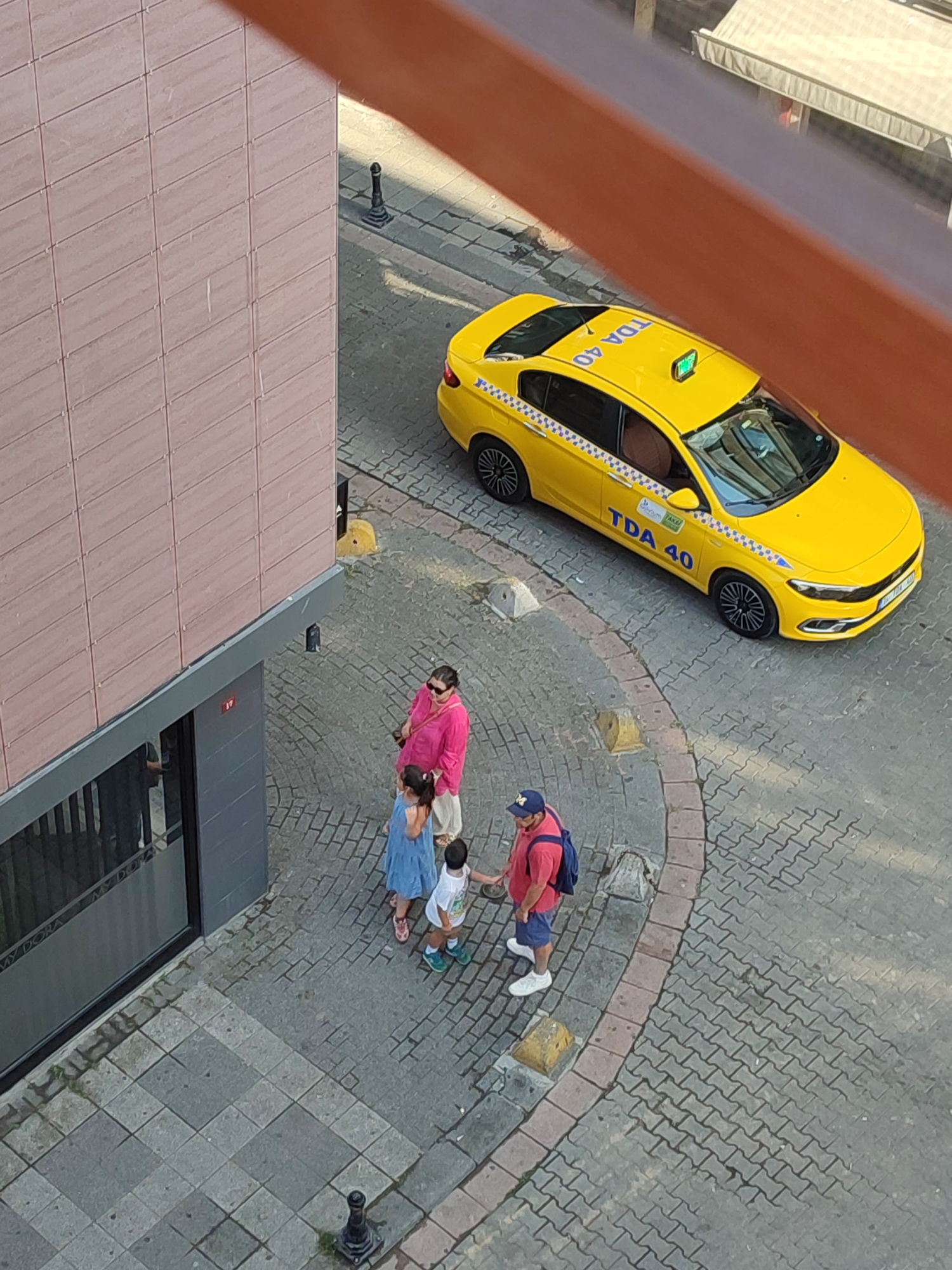 A yellow taxi with "TDA 40" visible on its roof is parked on a cobblestone street in front of a building. A family of four, two adults and two children, are standing on the sidewalk near the taxi. The adult female is wearing a pink shirt and sunglasses. The adult male is wearing a red shirt and blue shorts, and a blue backpack. A young girl in a blue dress and a young boy in a white shirt and blue shorts are standing between the adults. The building has a light pink tiled facade and a dark gray lower section with a window. The scene appears to be in a city, likely in Turkey given the taxi's markings and the presence of a lamp post.