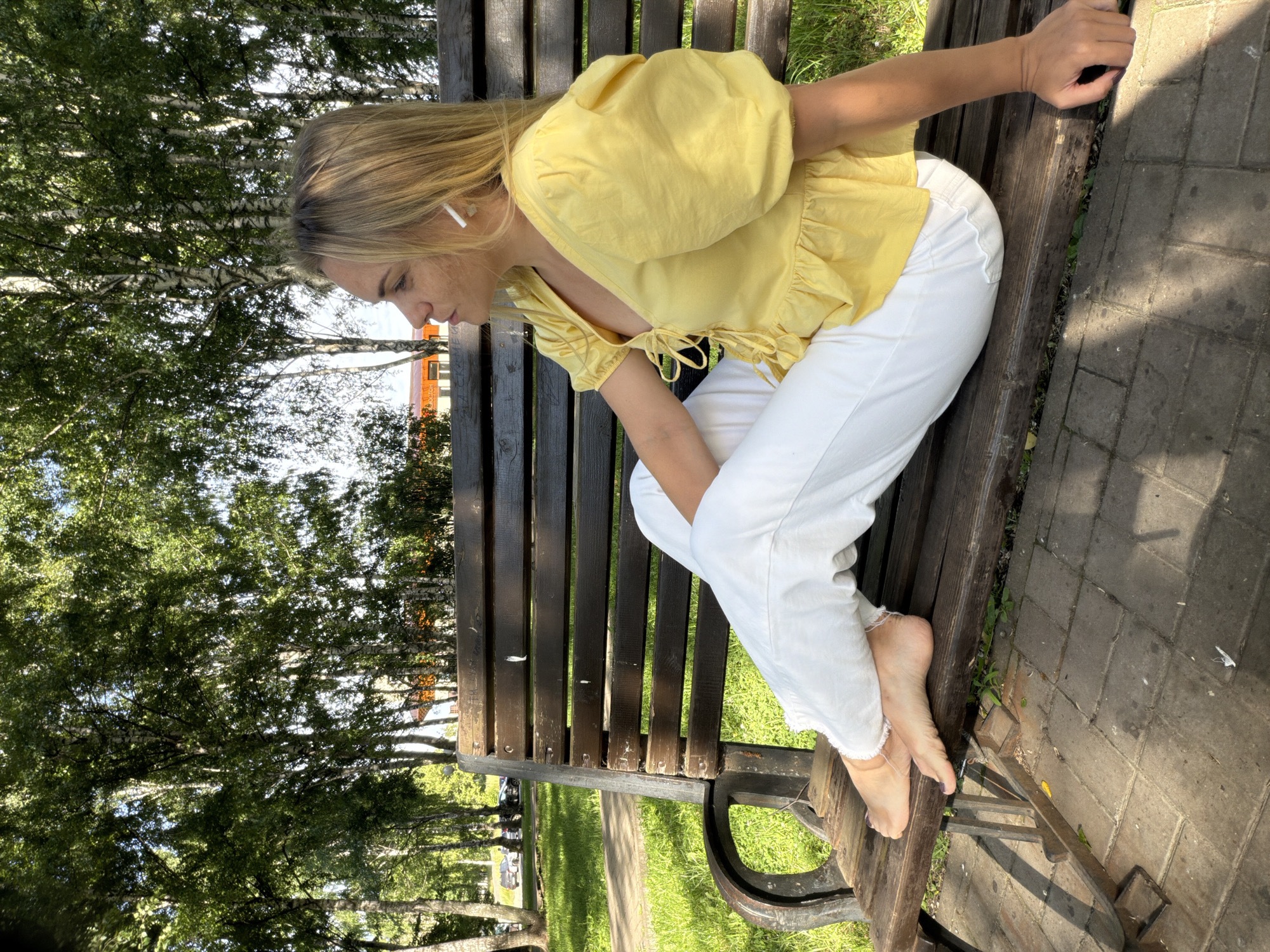 A young woman is sitting on a wooden park bench in Votkinsk, Russia. She is wearing a yellow puff-sleeved blouse and white pants. She has blonde hair and is wearing white wireless earbuds. She is looking down, with her head tilted to the side, and appears to be deep in thought. Her bare feet are resting on the bench. The bench is positioned in a park with trees and green grass. The sun is shining, casting shadows on the paved walkway next to the bench, indicating it is daytime. The overall mood seems peaceful and relaxed.