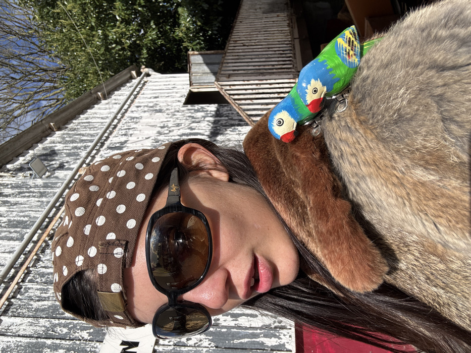 A close-up outdoor portrait in Neuilly-Plaisance, France, features a female subject positioned slightly right of center, looking towards the left with her mouth slightly open. She wears a brown fabric cap with white polka dots, worn backward, and large, dark, oval-shaped sunglasses with reflective brown-tinted lenses (Maui Jim branded). Her long dark hair is visible on her left side. She is dressed in a fur or faux fur coat, distinguished by a darker brown collar and a lighter, mottled brown body. Two small, brightly painted wooden parrot brooches, featuring blue heads, white faces with red beaks, and green bodies, are pinned to her right shoulder.
The background consists of a weathered wooden building with vertical planks, predominantly white with significant peeling paint revealing darker wood. A white outdoor light fixture is mounted on the building's wall. To the right, a wooden staircase and railing lead to an open doorway on an elevated section of the structure. Behind the building on the right, green tree foliage is visible against a clear blue sky, indicating strong natural sunlight.