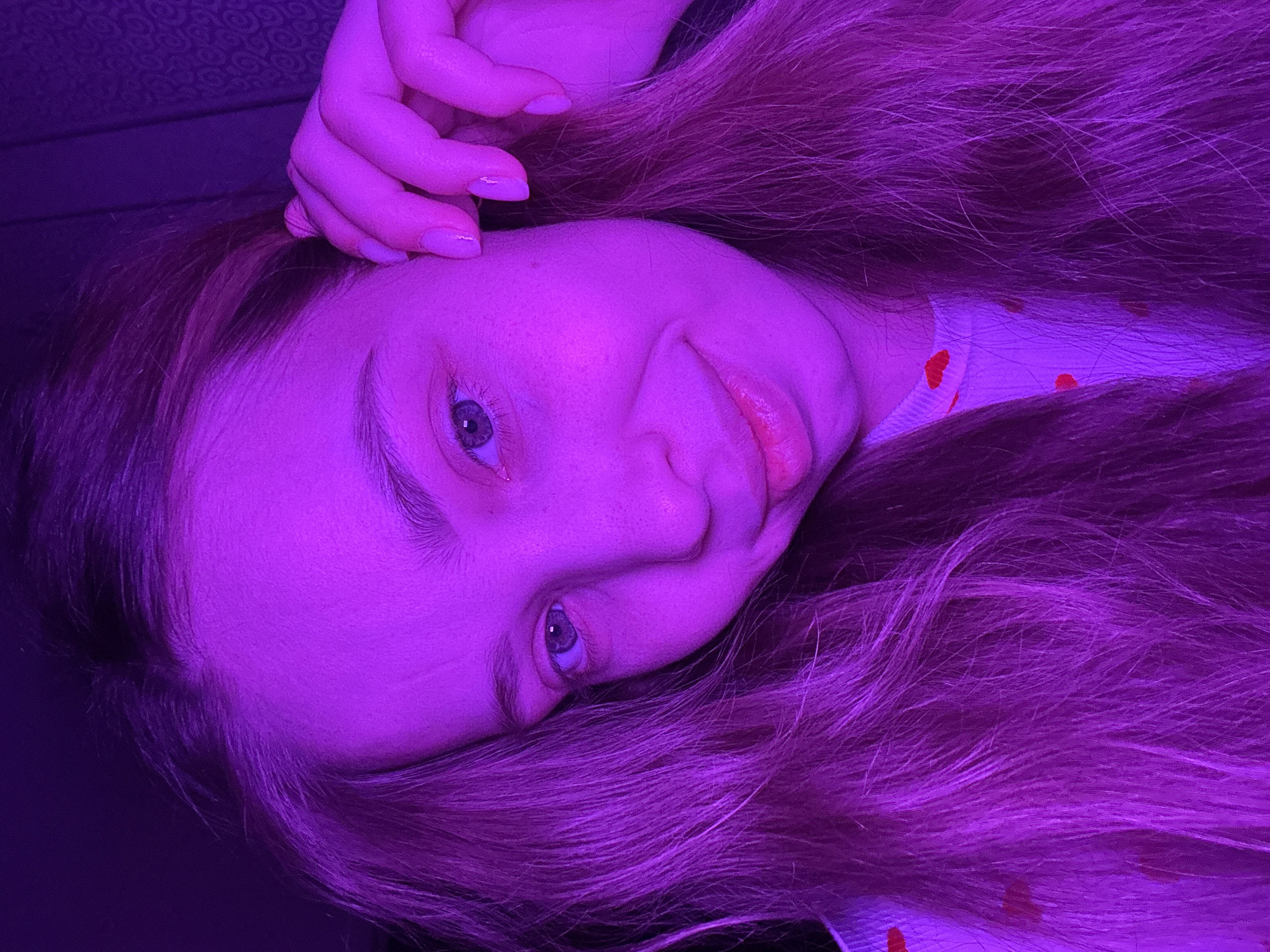 A close-up image displays a young female subject situated indoors within the city of Chaikovsky, Russia. The scene is illuminated by a strong magenta-purple light source, casting a monochromatic hue over the subject and her immediate background. The subject is positioned with her head slightly tilted, looking directly towards the camera with a subtle smile. Her right eye is fully visible, featuring a light-colored iris, and a portion of her left eye is also discernible. Her skin exhibits a fair complexion with scattered freckles visible on her cheeks. Long, light-colored hair, appearing purple under the lighting, frames her face and extends downwards. The subject's left hand is raised towards the left side of her head, with fingers gently touching her hair near her ear. Her fingernails are visible and appear manicured with a light-colored polish. She is wearing a light-colored garment, visible on her left shoulder and chest, adorned with a repeating pattern of small red hearts. The background is dark, with a faint, repeating pattern visible on a surface behind the subject's head to the right.