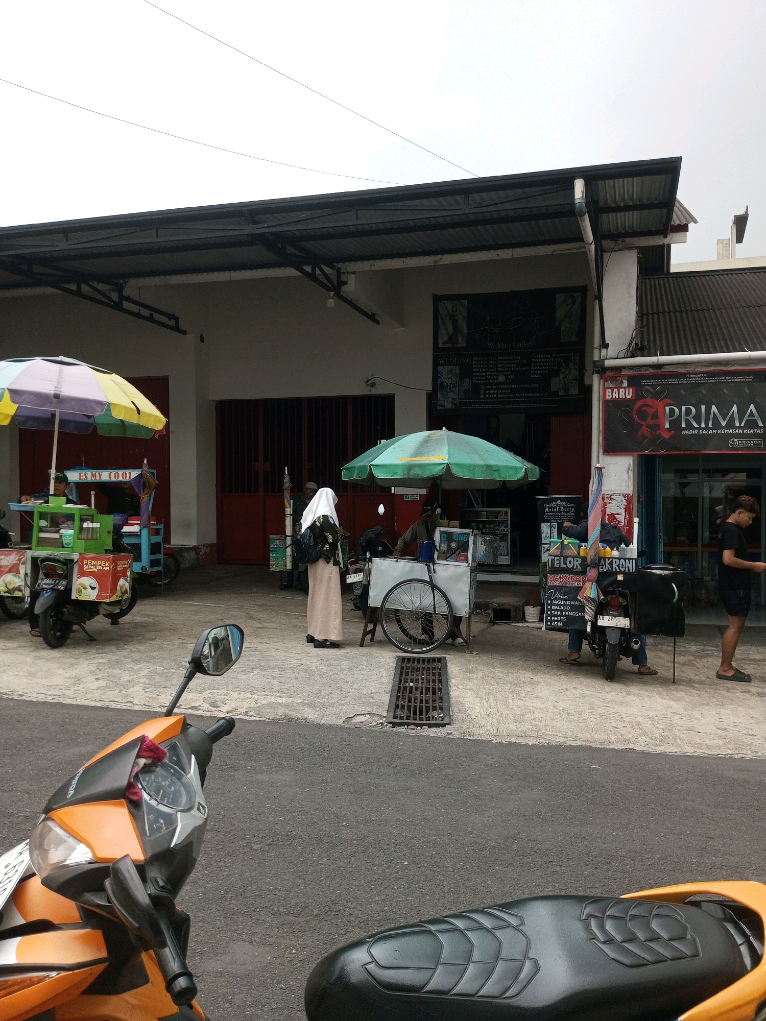 The image depicts a street scene in Krasak, Indonesia. In the foreground, an orange and black motorcycle is parked, its handlebars and front section visible. Behind it, the street transitions to a concrete walkway where several food vendors are set up.
To the left, a food cart with a multicolored umbrella sells what appears to be "ES MY COOL". A person is visible behind the counter. Adjacent to this cart is another, displaying a sign for "PEMPEK IKAN SELAM".
In the center of the scene, a woman wearing a hijab and a long, beige skirt stands near a green umbrella. Beside her is a food cart with a sign that reads "TELOR MAKRONI AKRONI" and other menu items. A motorcycle is attached to this cart.
To the right, a man wearing a black t-shirt and shorts stands near the entrance of a building. A sign above the entrance advertises "BARU PRIMA". The building appears to be a shop or business.
The environment suggests an outdoor marketplace or a street with multiple small businesses. The sky is overcast.