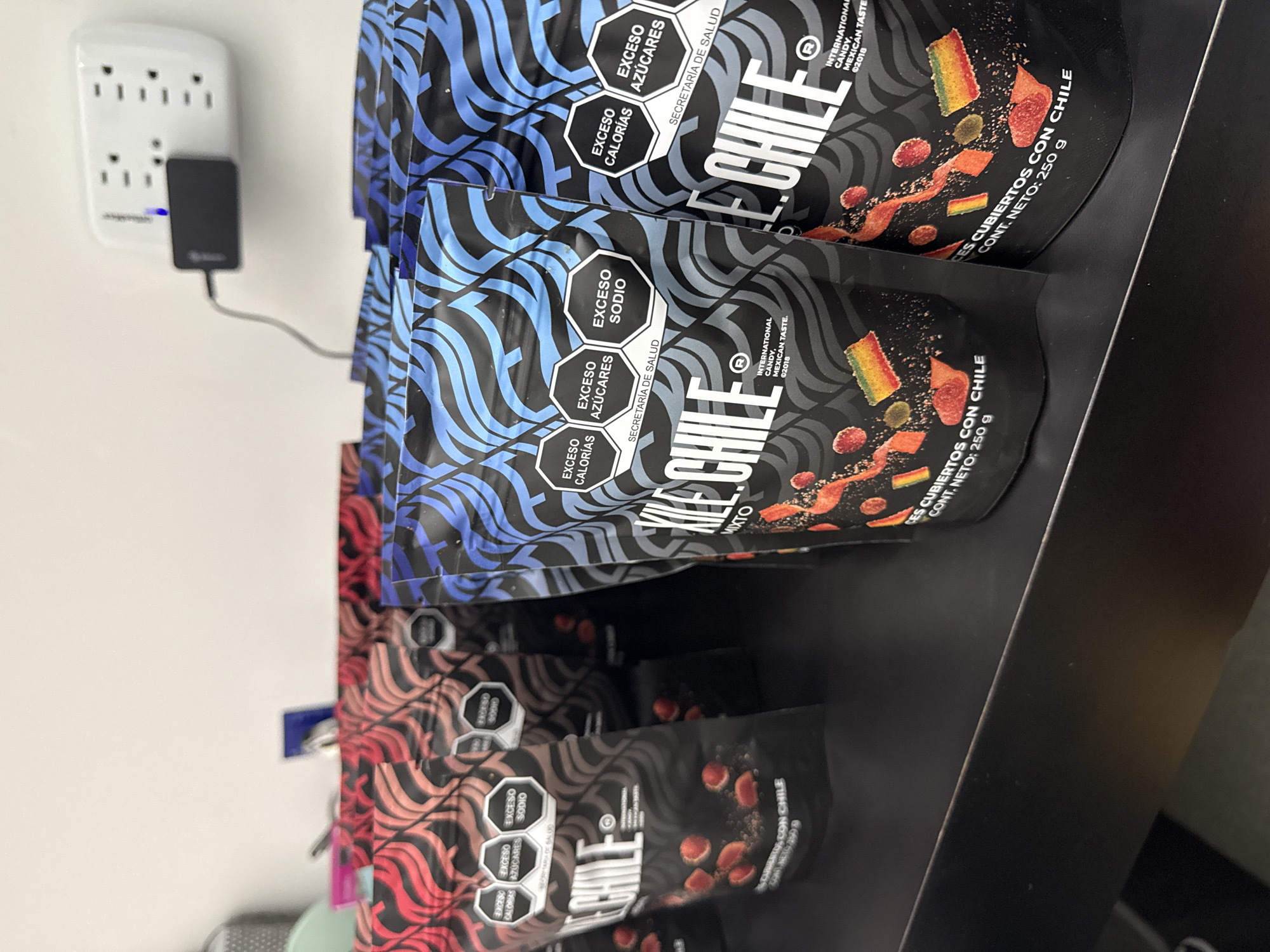 The image shows a shelf of candy bags with a power outlet visible on the wall. The candy, labeled "LE CHILE" in large letters, is in black packaging with a wavy pattern design in different color variations: blue, and red. Each bag has a hexagonal warning label in Spanish indicating "EXCESO CALORIAS" (excess calories), "EXCESO AZUCARES" (excess sugars), and "EXCESO SODIO" (excess sodium). The text "INTERNATIONAL CANDY, MEXICAN TASTE" is printed in smaller letters with a 2018 copyright date. The label also reads "CES CUBIERTOS CON CHILE CONT. NETO: 250 g", indicating that the bag contains candy covered with chili and the net weight is 250 grams. A black power adapter is plugged into the power outlet on the wall. The location is indeterminate.