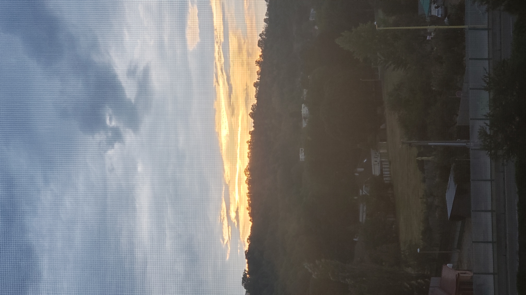 The image depicts a landscape view, likely captured during sunset or sunrise, through a window with a visible screen texture. A silhouetted hill covered in trees dominates the middle ground, with a band of bright, warm light illuminating the sky above its crest. A few buildings with light-colored walls and darker roofs are scattered on the hillside. The foreground shows a road or pathway with a grey barrier or fence running horizontally. Various structures, including a small red-roofed building, a dark shed, and a yellow lamppost, are visible in the lower portion of the image. The sky is a mix of grey and white clouds, with streaks of yellow and orange light indicating the sun's position. The overall impression is one of a semi-rural or suburban setting on the outskirts of a city. While the image does not explicitly confirm the location, it is stated to be in Prešov, Slovakia.