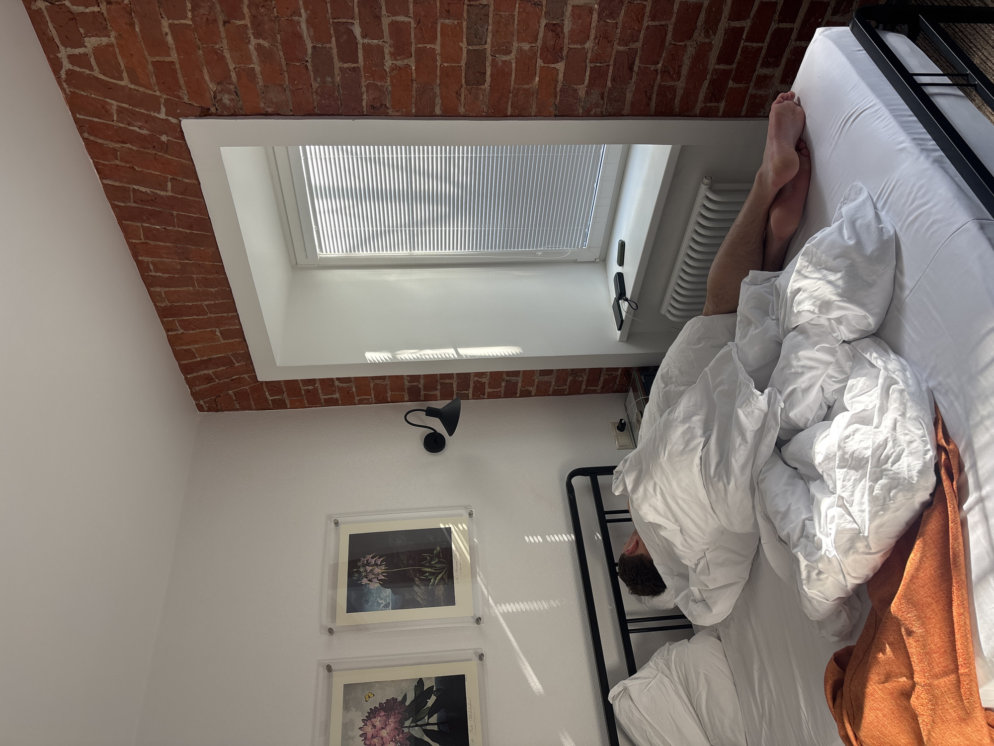The image shows a person sleeping in a bed by a window. The bed has white sheets and a black metal frame. An orange blanket is partially covering the bed. The person's legs are visible, resting near the window. The window has white blinds and is set into a wall of exposed red brick. The room has white walls and two framed botanical prints hanging on the wall. A black adjustable lamp is also mounted on the wall. The location is likely a residential interior, potentially an apartment in Moscow, Russia, given the architectural style of the brickwork.
