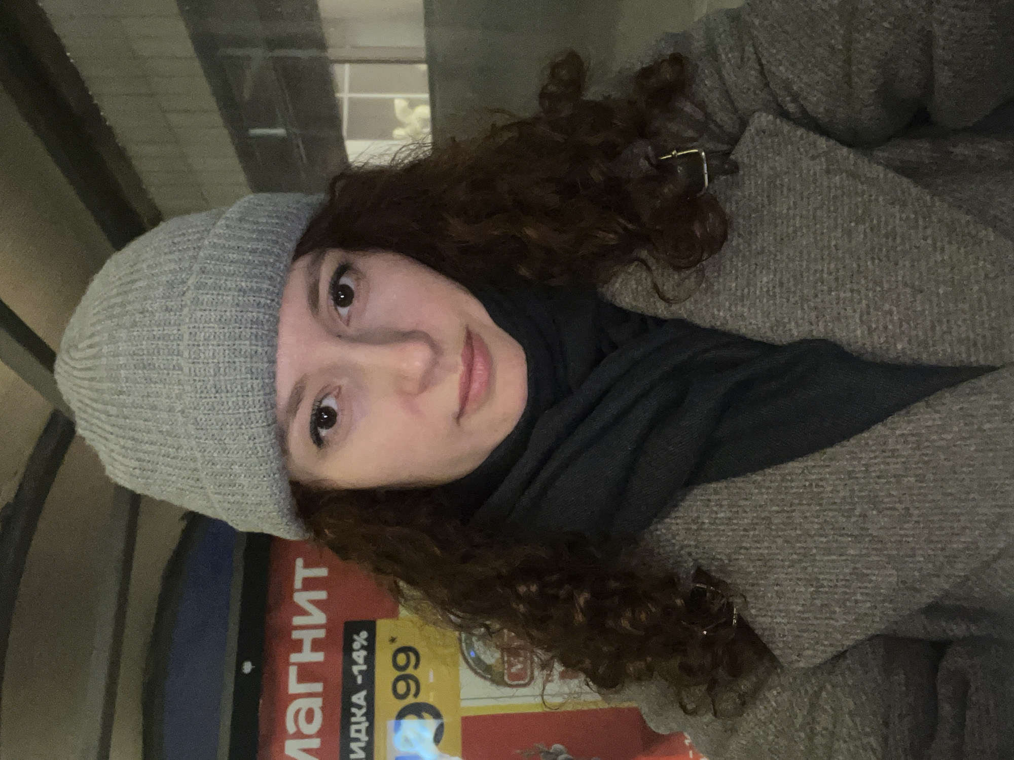 A woman with curly, reddish-brown hair wearing a gray beanie, a dark gray scarf, and a gray tweed coat is posing for a selfie. She is standing in front of an advertisement for a "Magnit" store, which features Russian text and prices. The text indicates the store is advertising vodka at a 14% discount. The background suggests an urban environment, possibly a bus stop or an outdoor shopping area. The location is most likely in Russia, based on the store and the Russian text in the advertisement. Given the presence of a "Magnit" store, the location could be Saint Petersburg, Russia.