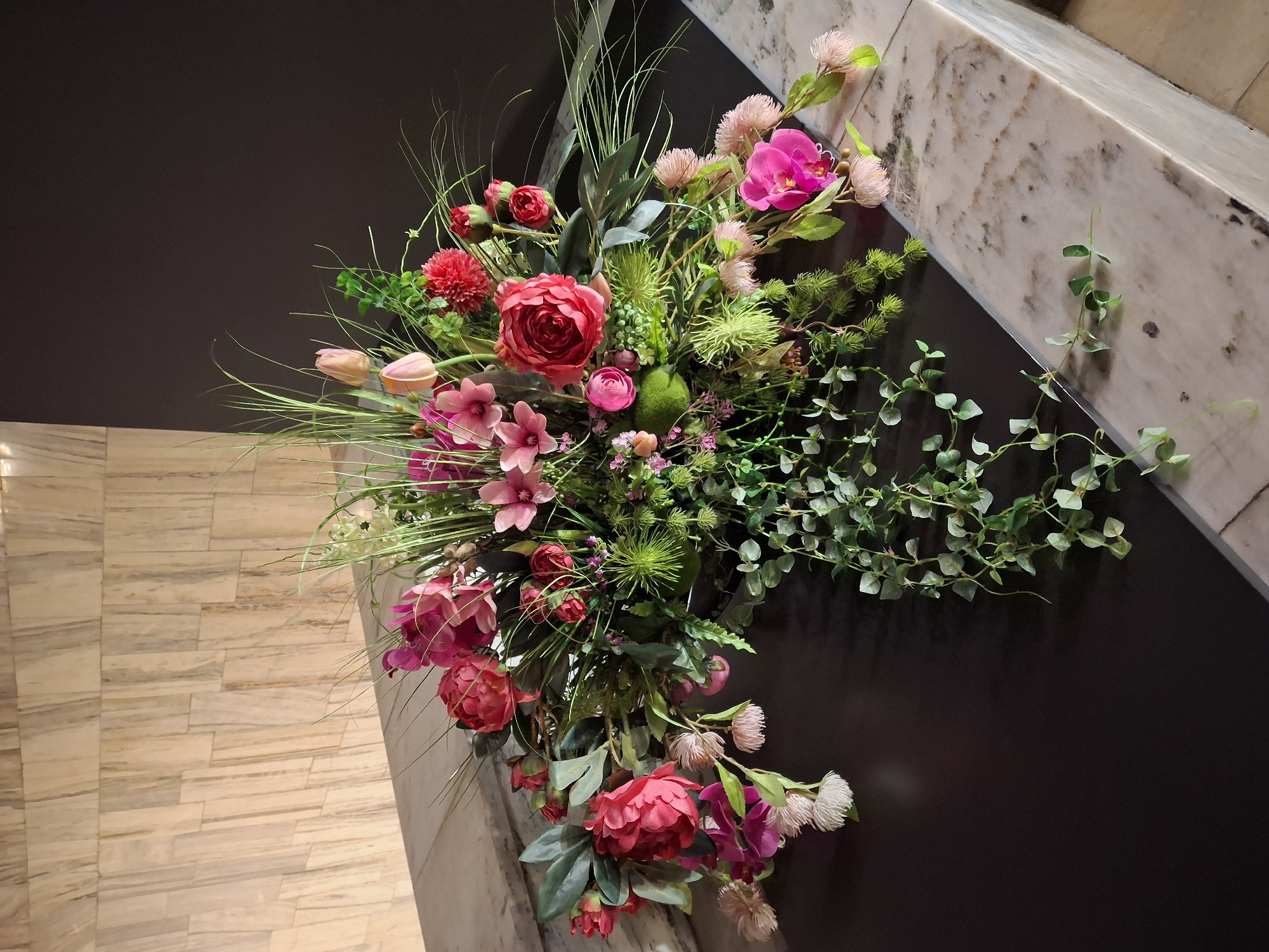The image shows a floral arrangement on a dark surface, possibly a countertop or table, with a marble edge. Behind the arrangement is a wall with a vertical patterned texture, likely stone or tile. The floral arrangement is lush and features a variety of flowers in shades of pink, red, and fuchsia, along with greenery and wispy grass-like elements. Several pink tulips are visible, as are large peony-like flowers, smaller pink blossoms, and a bright pink orchid. A cascading vine with small green leaves trails down the side of the arrangement. The scene is well-lit, casting soft shadows. The location is described as being in the city of Pila, Poland, within a building.