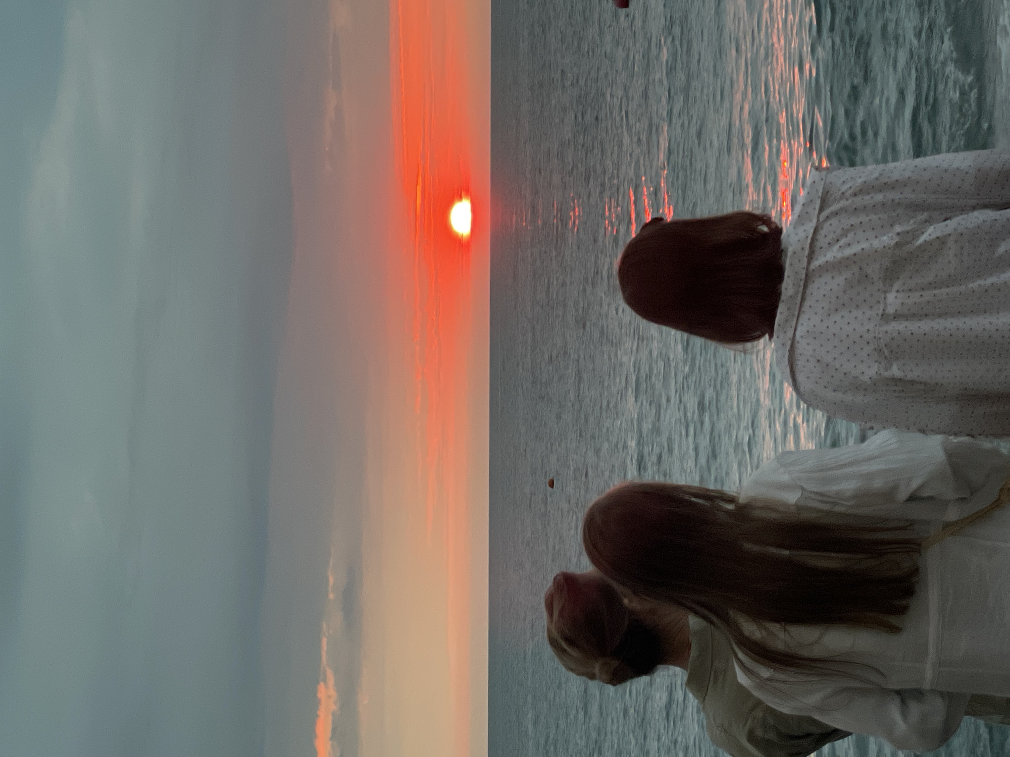 The image depicts three individuals facing away from the camera, observing a vibrant sunset over the Black Sea. The sky is a gradient of muted gray and pink, with the sun appearing as a bright, fiery orb on the horizon, casting a shimmering reflection on the water. The sea itself is a deep blue-green, with gentle waves. The people are standing on what appears to be a promenade or pier. The person on the far left has their hair tied back and wears a light-colored shirt. The person in the middle has long, dark hair and wears a white blouse. The person on the right has short, dark hair and wears a white patterned shirt. The overall atmosphere is one of tranquil observation of the natural beauty of the sunset. This scene is consistent with coastal areas of Sochi, Russia, known for its Black Sea coastline and dramatic sunsets.