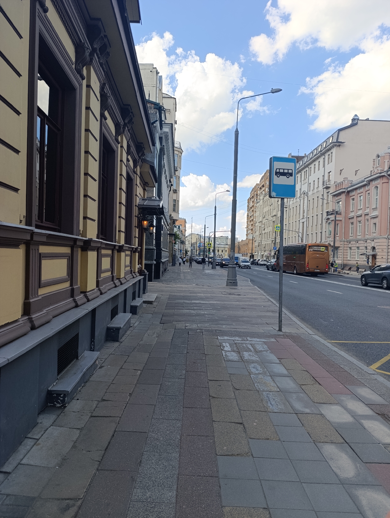 This media file depicts a daytime street scene in Moscow, Russia, viewed from a sidewalk looking down a long, urban thoroughfare. The sky above is bright blue with scattered white cumulus clouds, suggesting fair weather.
On the immediate left, a grand multi-story building with a light-yellow facade and dark brown architectural embellishments dominates the foreground. These details include ornate window frames, decorative corbels under the eaves, and pilasters along the wall. An antique-style street lamp is affixed to its facade. The sidewalk below is paved with a mosaic of rectangular tiles in varying shades of grey and reddish-brown, some appearing worn or mismatched.
The street extends into the distance, flanked by a continuous line of multi-story buildings that display a mix of architectural styles. On the right side of the image, the road features vehicle traffic. A large, brown-orange bus is prominently seen driving away from what appears to be a bus stop. A blue square bus stop sign, featuring a white bus pictogram, is centrally located on a pole beside the sidewalk, marking the stop. Further down the street, additional traffic signs are visible, including a yellow diamond-shaped pedestrian crossing sign and a blue 'P' sign for parking. Several cars are present on the road, both moving and parked.
A few pedestrians can be observed walking on the sidewalks in the distance. The scene captures the routine activity of an urban setting during daylight. Any text on the back of the brown bus is too small to be legible.