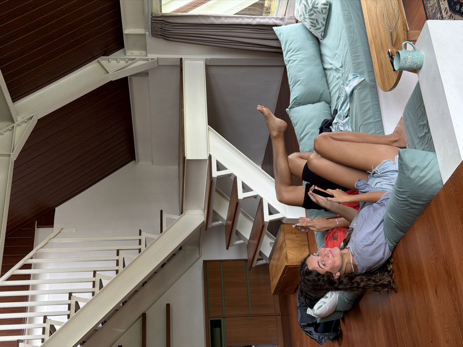 A woman with long, curly hair, wearing a gray shirt, lies on a light blue couch. Her legs are intertwined with a person wearing black shorts and the two are looking at a phone. The scene appears to be inside a modern house with a wooden floor and white staircase with light brown steps, visible in the background. A brown ceiling is partially visible on the upper left.