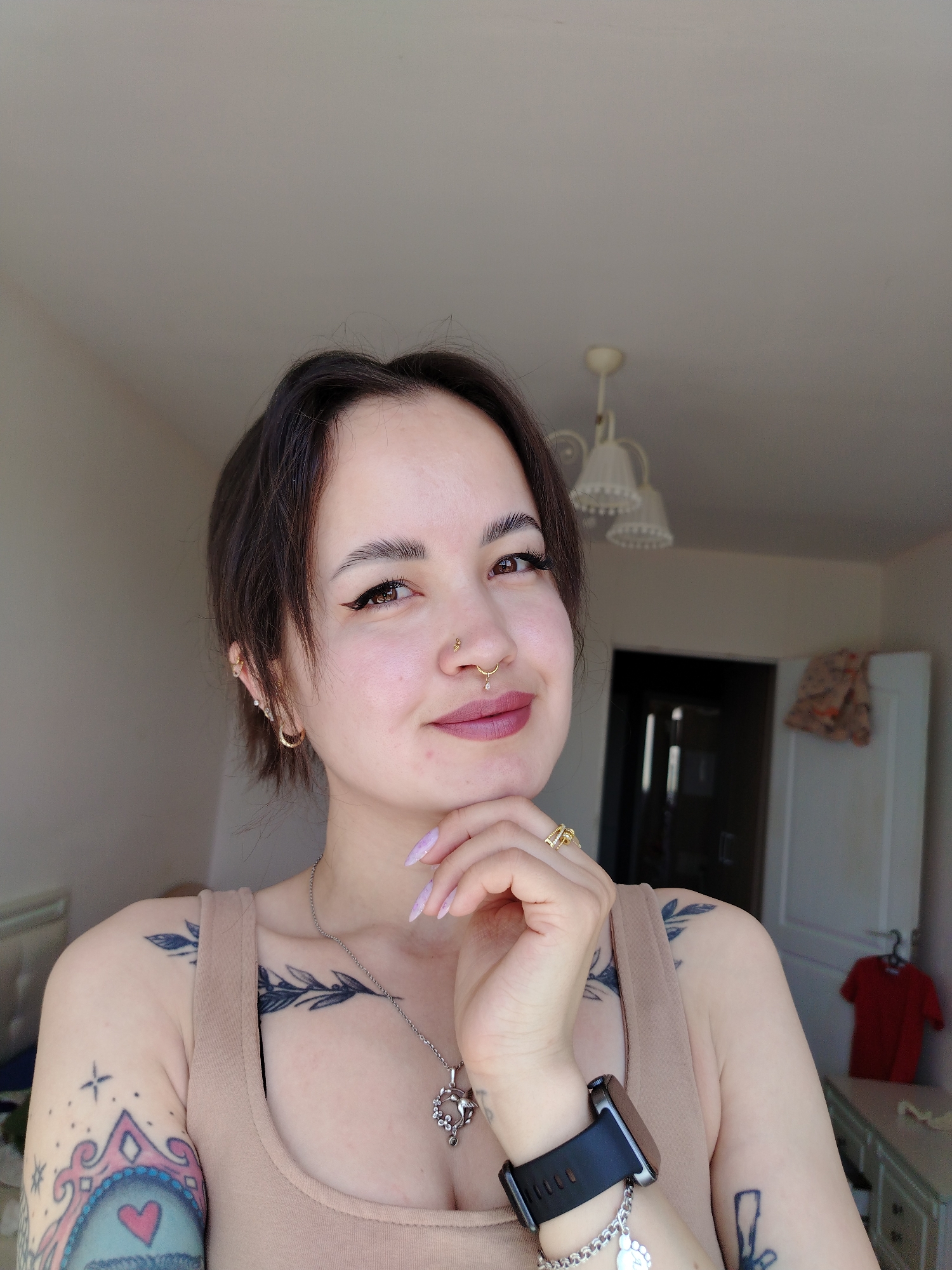 A woman is indoors with a light-colored wall behind her. She has dark hair and eyebrows, light skin, and brown eyes. She is wearing a tan tank top, a necklace, and multiple rings. She has numerous tattoos on her arms, chest, and shoulders. She is wearing a smartwatch and has multiple piercings in her ears and nose. Her hand is near her chin, and she is looking towards the viewer with a slight smile. In the background, there is a door, a dresser, a red t-shirt hanging on a hanger, and a light fixture on the ceiling. The location is unknown, and there is no indication it is Noviy, Russia.