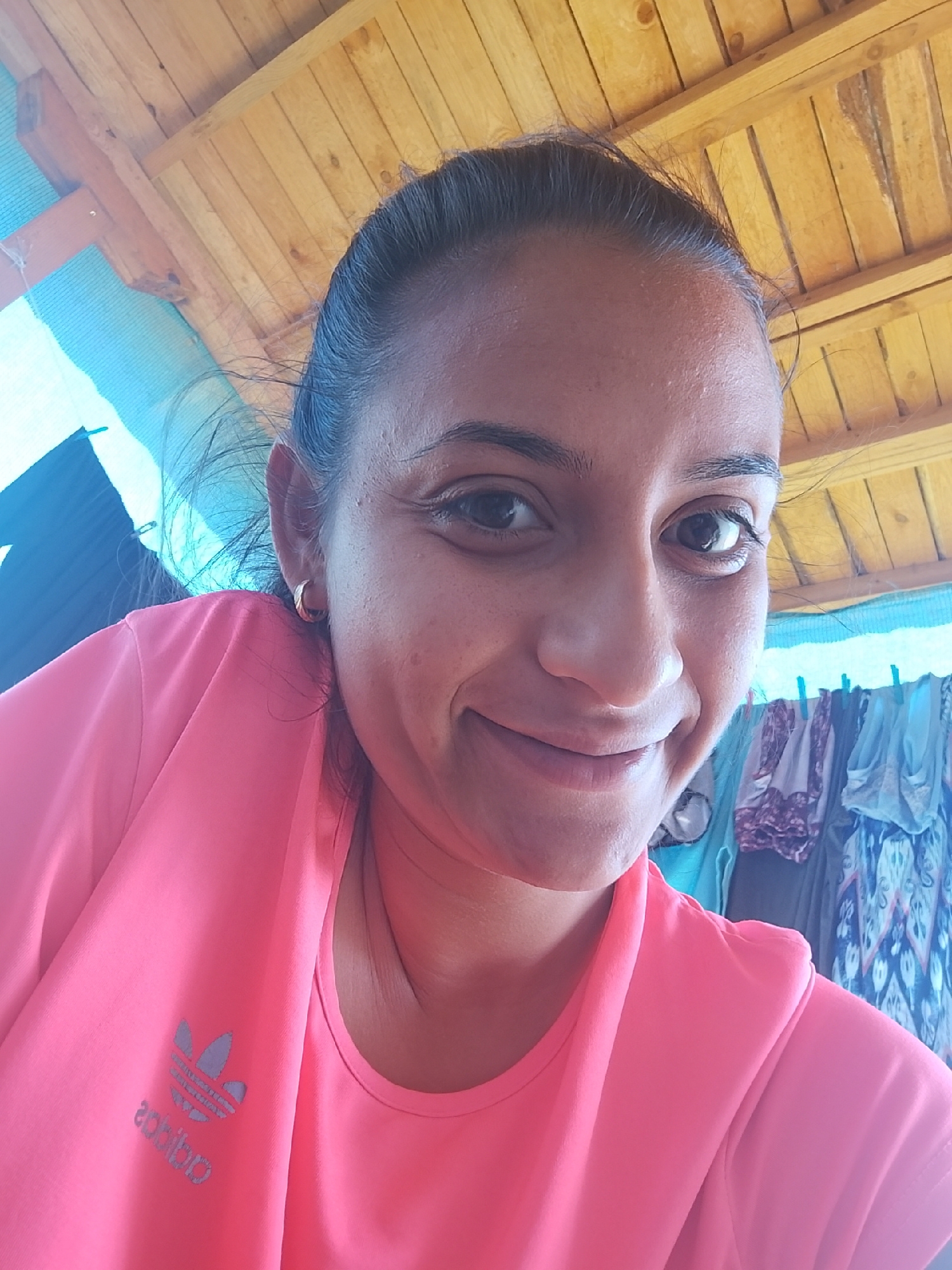 A woman with dark hair pulled back smiles at the camera, her head tilted slightly to her right. She is wearing a bright pink t-shirt with a reflective Adidas logo on the left chest. The background is a rustic wooden ceiling, with hints of blue netting and what appears to be laundry hanging to her right. The lighting suggests an outdoor, possibly covered, area. The image was taken in Zimnitsa, Bulgaria.