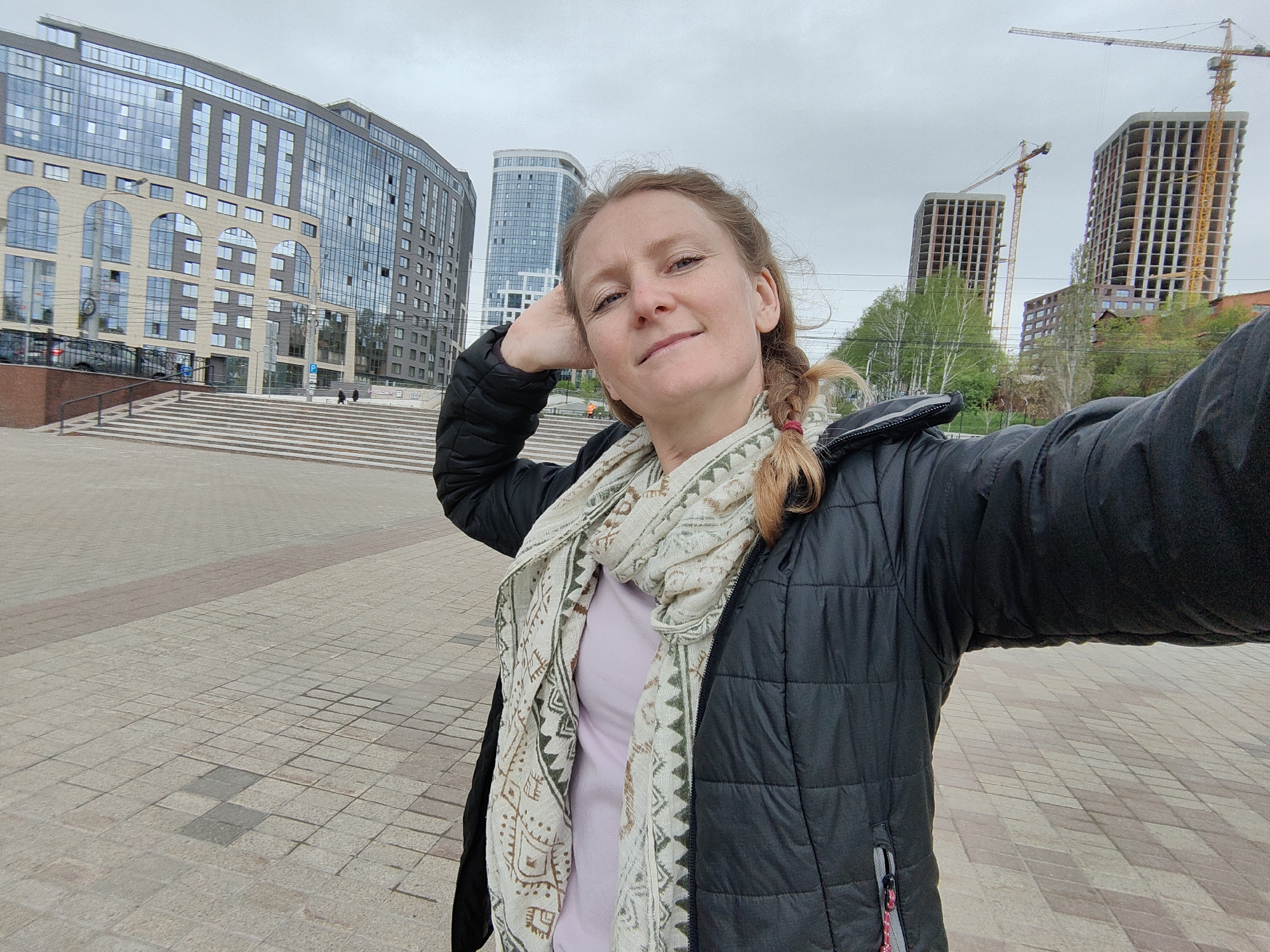 The image is a selfie of a woman, likely in her 30s or 40s, taken outdoors in Izhevsk, Russia. She is wearing a black puffy jacket and a patterned scarf. Her hair is in a braid. In the background, there are modern buildings and structures, including some under construction with visible cranes. The ground appears to be paved with cobblestones. The weather seems overcast. The woman is positioned in the foreground, with the city buildings forming the backdrop.