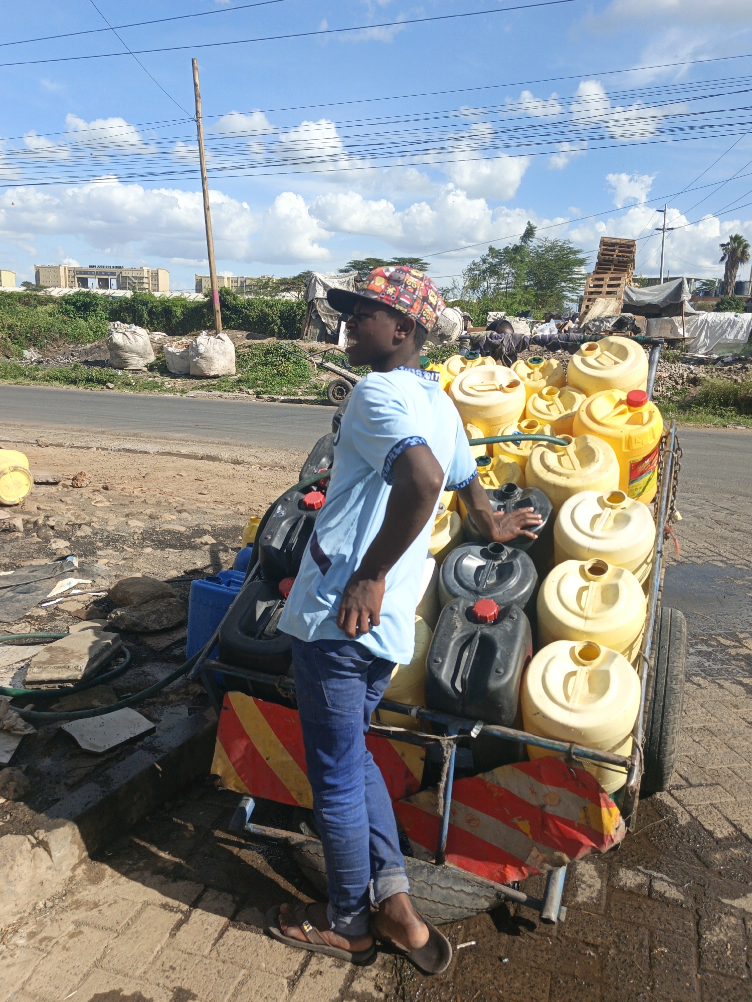 A man stands next to a cart laden with yellow and black plastic water containers on a street in Nairobi, Kenya. The man wears a colorful cap, light blue shirt, jeans, and sandals. He rests his left hand on a container. The cart has a striped red and white warning panel on the back. A hose connects to a blue container on the ground next to the cart, indicating water transfer. The background shows a road, buildings, bags of waste, and a structure made of wooden pallets, reflecting an urban environment.