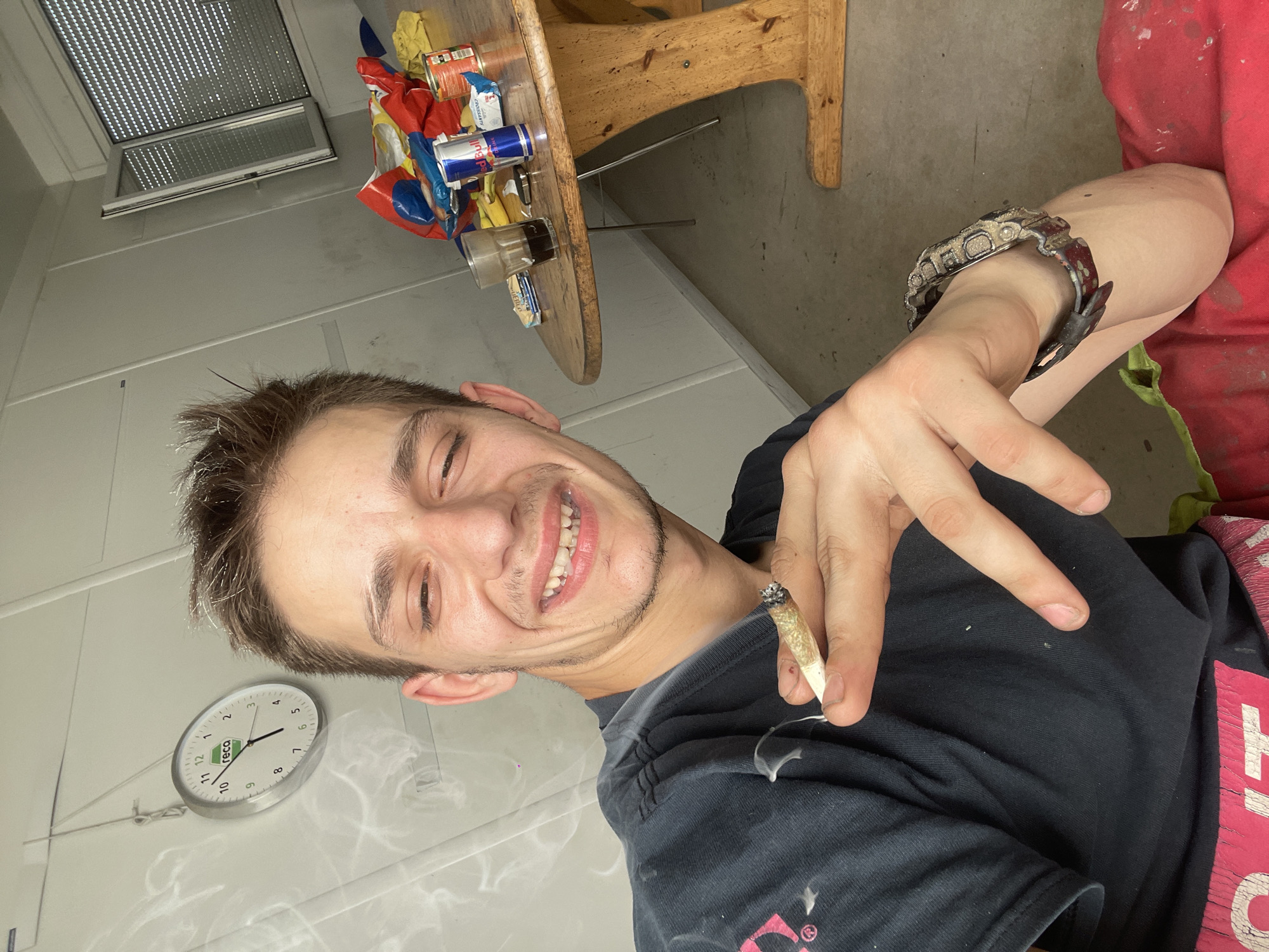 A medium shot captures a young Caucasian male with short, light brown hair, a light stubble, and a wide smile as he holds a lit cigarette between his index and middle fingers. He is wearing a dark t-shirt with a pink graphic design on the left side. The background includes a white wall with a round clock showing approximately 10:48, a table laden with snacks, energy drinks, and a glass of dark liquid, and a partially open window with blinds. The room appears to be an indoor space, possibly a break room or workshop, with a concrete floor. The lighting suggests an indoor environment with natural light coming from the window.