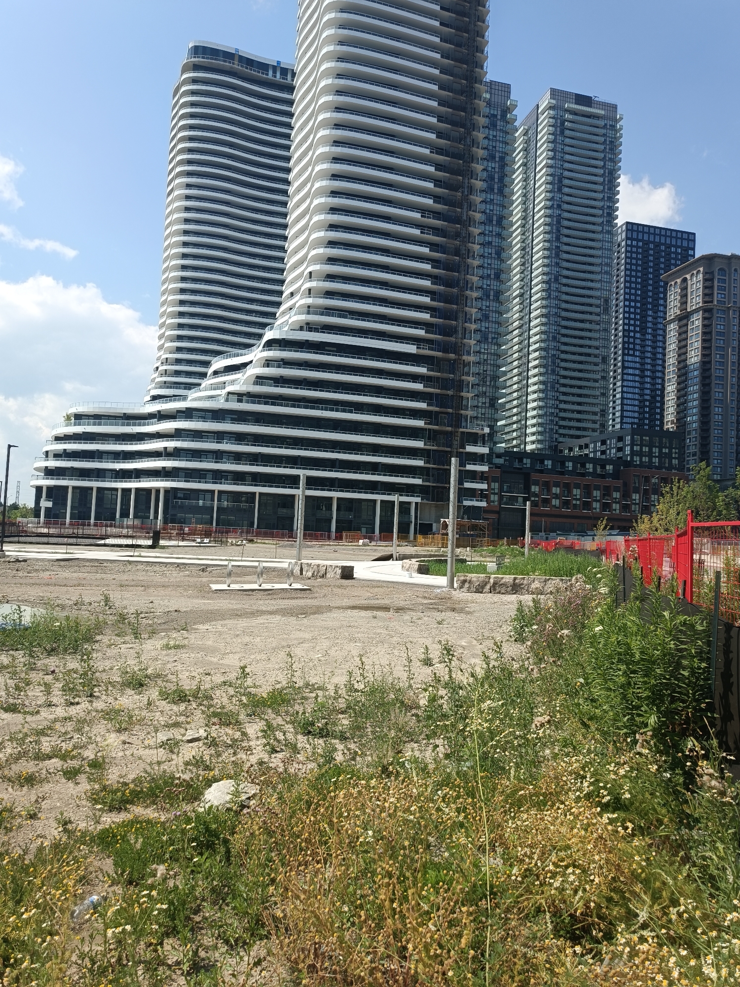 This is a landscape photograph of a construction site in Mississauga, Canada, with several modern skyscrapers in the background. In the foreground, there is a patch of dry, sandy ground covered with weeds and some scattered rocks. A plastic bottle lies on the ground in the lower left corner, partially obscured by vegetation. A red fence runs along the right side of the image, separating the construction area from another part of the property. Behind the fence, more greenery and the side of a dark building are visible.
The skyscrapers in the background are predominantly glass and white, with curved balconies creating a wave-like effect on some of the buildings. One particularly striking building features a distinctive undulating white facade. Other buildings are more traditional rectangular towers with visible rows of balconies. The sky is a clear blue with a few scattered white clouds. In the mid-ground, there is a paved walkway with some metal bike racks and large stones placed as decorative elements. Several construction cranes are visible in the distance, indicating ongoing development. The scene appears to be a bright, sunny day.