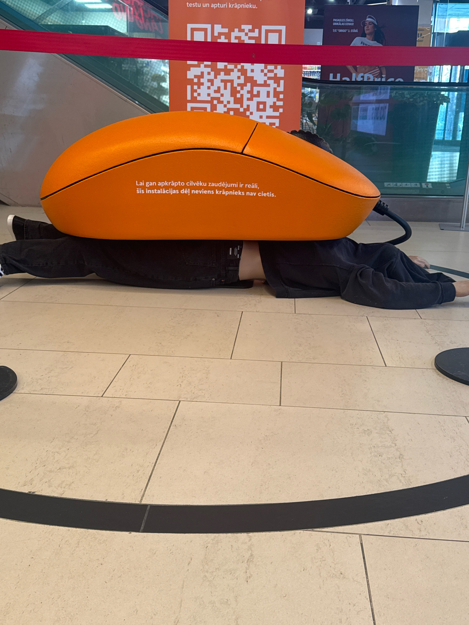 The image shows a person lying on their back on a tiled floor, with a large, orange, computer mouse-shaped object on top of them. The person is wearing black jeans and a black long-sleeved shirt. Their legs are extended and their arms are relaxed. The orange mouse is positioned with its base on the person's torso and legs. Text is visible on the orange mouse, written in Latvian, which reads: "Lai gan apkrāpto cilvēku zaudējumi ir reāli, šis instalācijas dēļ neviens krāpnieks nav cietis." This translates to: "Although the losses of the cheated people are real, no fraudster has suffered because of this installation."
In the background, there is a red ribbon barrier, a QR code, and a large advertisement featuring a woman. Behind the advertisement, there is a glass-enclosed escalator or moving walkway and glimpses of a retail environment, possibly a store. The tiled floor has a pattern of light-colored square tiles with dark grout lines. A curved black line is also visible on the floor in the foreground. The scene appears to be indoors, likely in a public space such as a shopping mall or a public venue. Given the Latvian text on the object, the location is likely in Riga, Latvia.