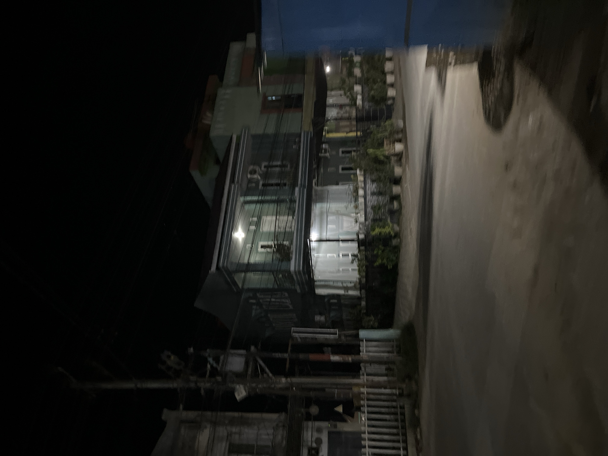 A nighttime street view in Landuh, Indonesia, shows a two-story building with green and gray siding. The building has a balcony on the second floor and is illuminated by interior lights. Several potted plants are arranged along the front of the building. To the left, a white fence borders a sidewalk and a utility pole with wires. A blue structure is visible on the right edge of the frame. The street in the foreground is paved and appears wet, reflecting ambient light. The overall scene depicts a quiet urban residential or commercial area at night.