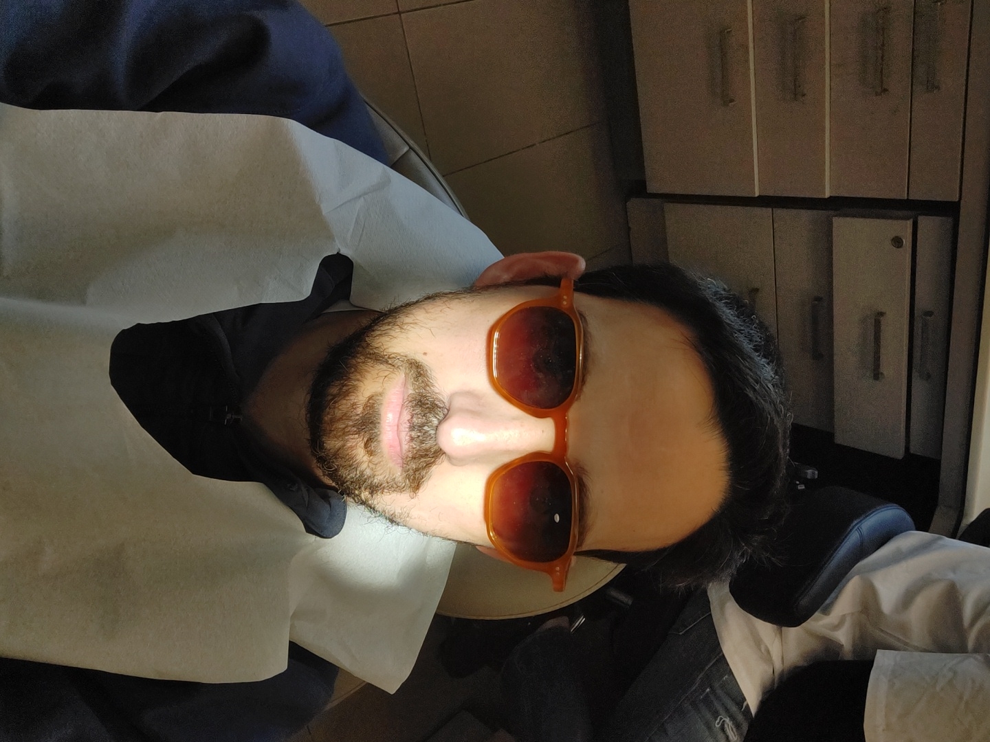 A bearded man wearing orange sunglasses is lying in what appears to be a dental or medical chair covered with a white paper sheet. The background shows a cabinet with several drawers, suggesting a clinic or office environment. There is not enough information to determine the specific location in Tel-Aviv, Israel.