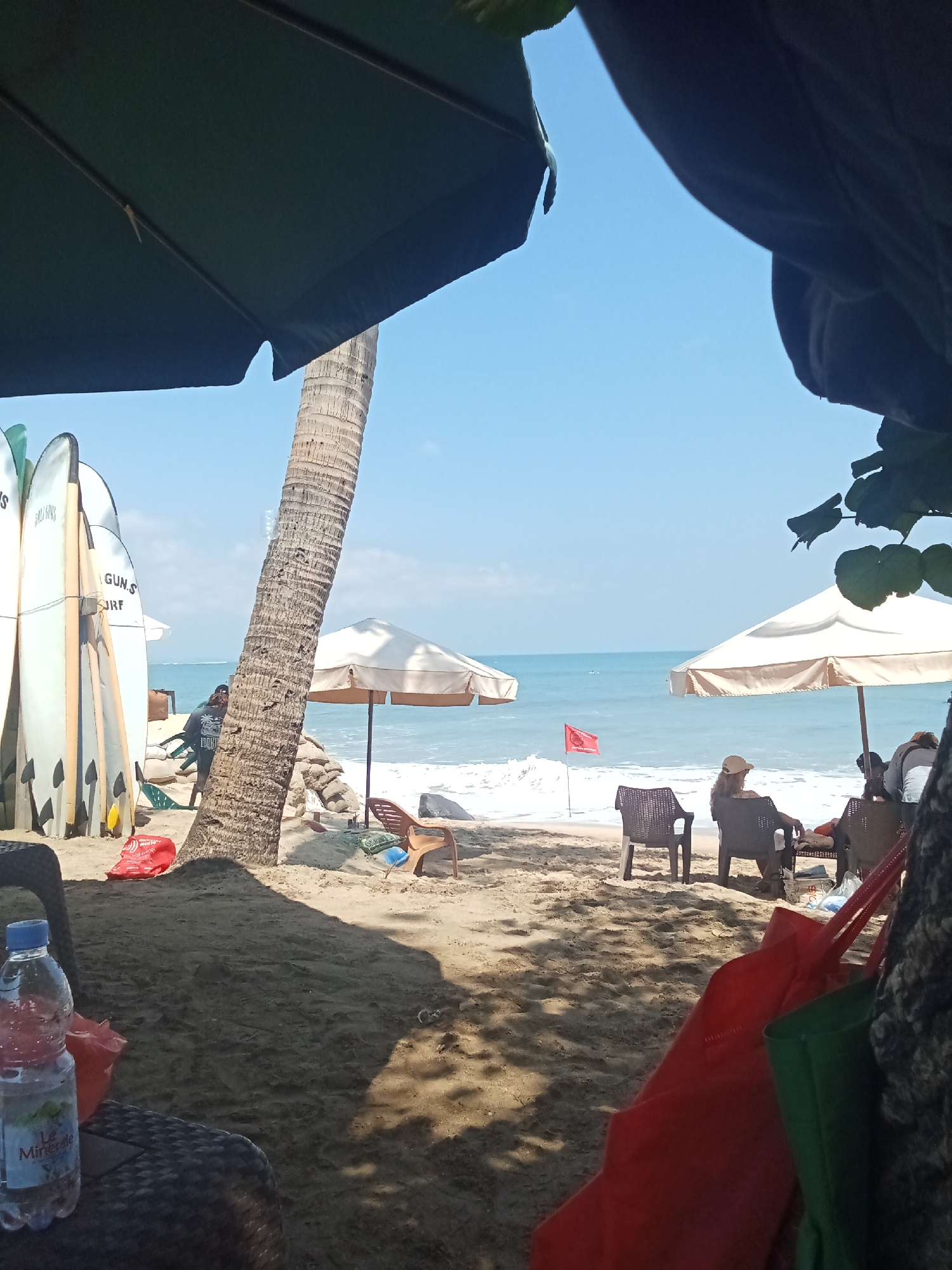 The image depicts a sunny day at a beach in Kuta, Indonesia. In the foreground, there is a bottle of water and two large tote bags, one red and one green, resting on a woven chair. The beach is sandy, with some shadows cast by umbrellas. To the left, several surfboards are propped up. A tall palm tree stands near the center of the frame. In the middle ground, two beach umbrellas are set up, providing shade for beach chairs. A few people are seated in these chairs, facing the ocean. The ocean is a vibrant blue with gentle waves. A red flag is planted in the sand near the water's edge, possibly indicating swimming conditions. In the background, the horizon shows a clear blue sky.