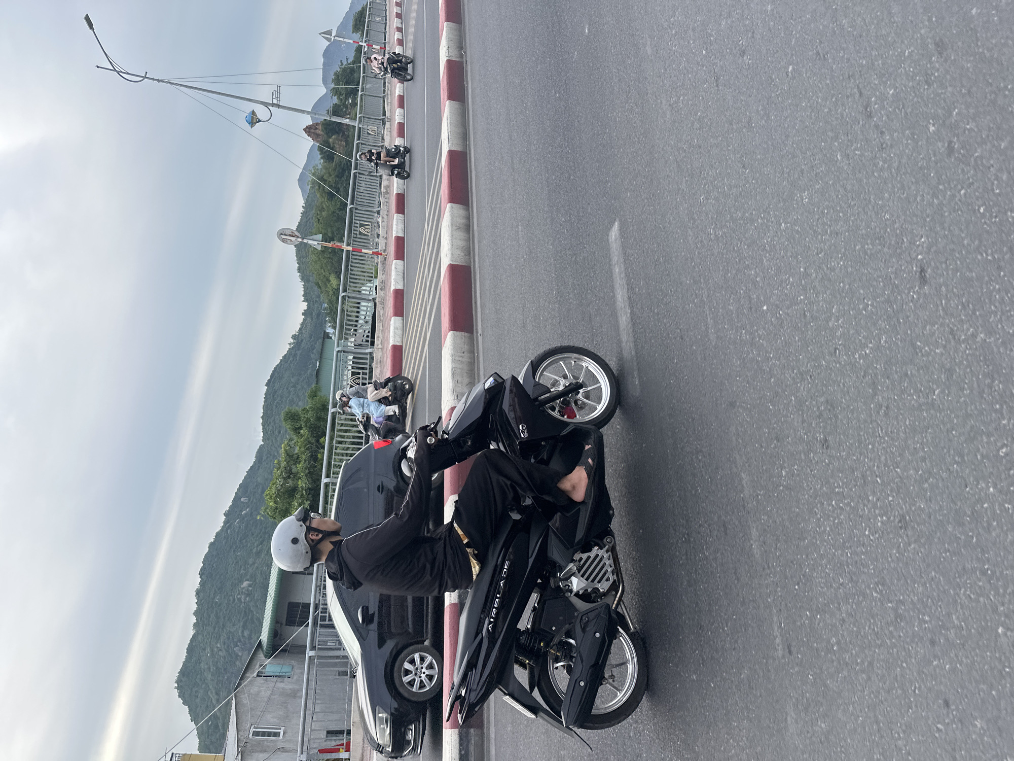 A man wearing a helmet and a black hoodie rides a black scooter on a road in Bac Nha Trang Ward, Vietnam. The scooter has the word "AIRBLADE" written on its side. The man is wearing dark pants and sandals. In the background, there are other people on scooters, a black car, a bridge with a red and white striped railing, trees, and mountains under a cloudy sky. The road surface is asphalt.