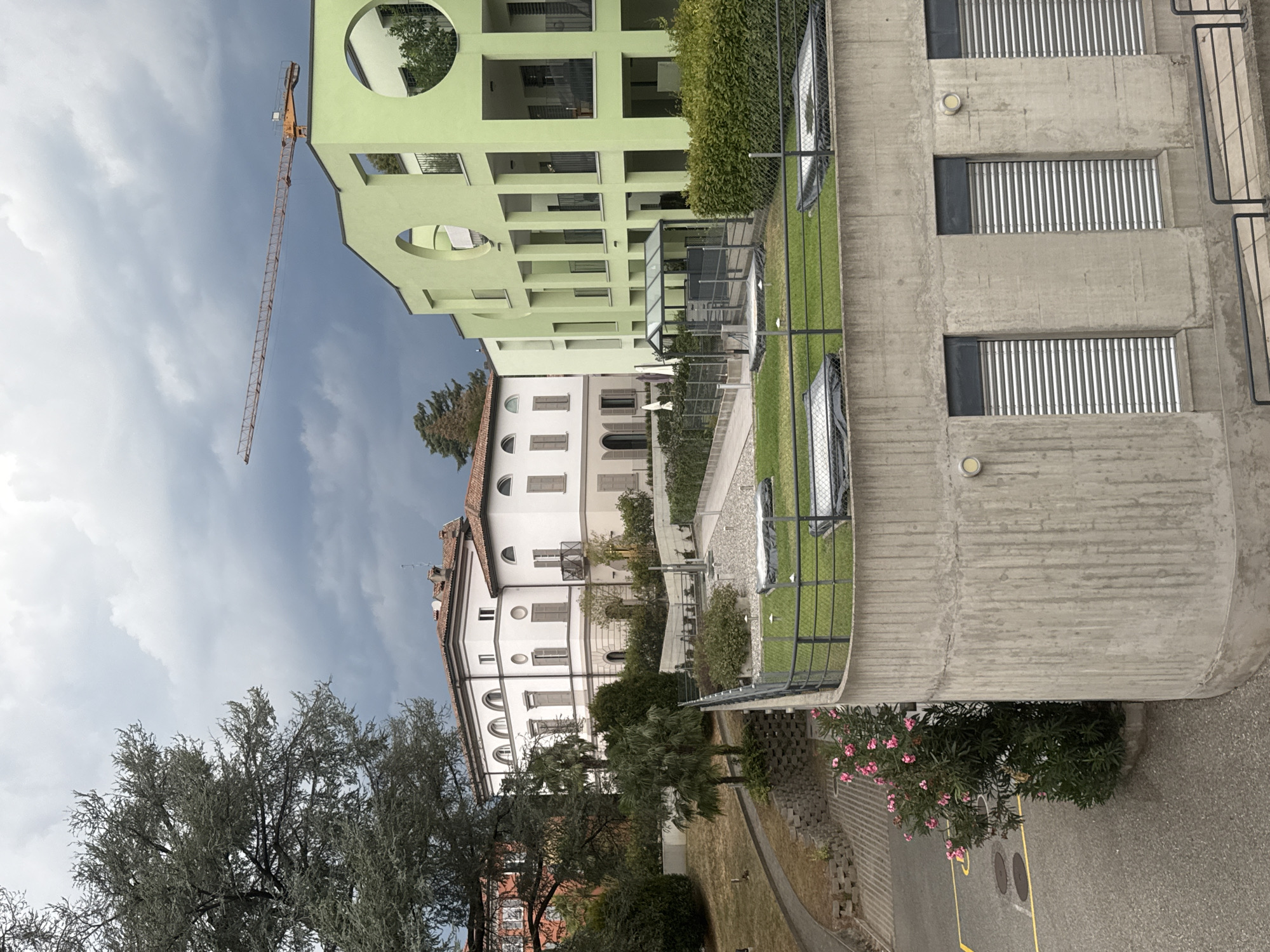 In Balerna, Switzerland, on a cloudy day, a complex of modern buildings is depicted. To the right, a lime green, multi-story building features a facade with large circular cutouts and recessed balconies. Adjacent to it is a white, classical-style building with arched windows and a red tiled roof. A construction crane with an orange boom extends horizontally across the sky above the green building. In the foreground, a concrete structure with horizontal blinds is visible, topped with a fenced-off area containing a green lawn and covered objects. A large tree with sparse, gray-green foliage stands to the left, partially obscuring the view of the buildings. A pathway, a patch of dry grass, and a blooming pink bush are also present. The scene appears to be an urban or suburban environment with elements of both historic and contemporary architecture, set against a dramatic, overcast sky.