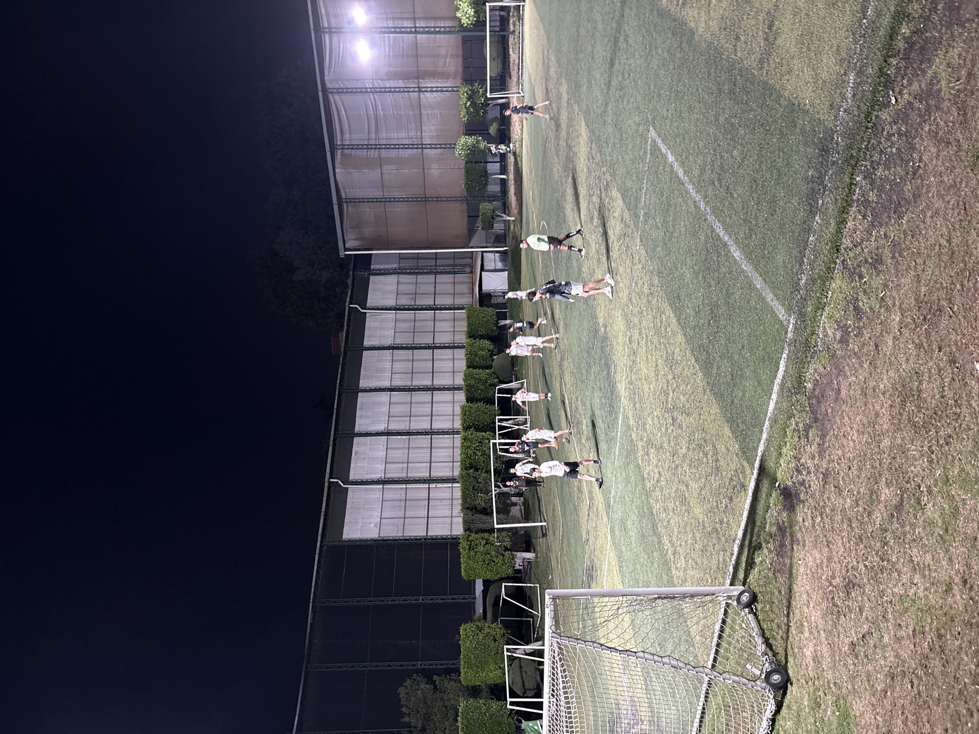 A brightly lit soccer field in Huixquilucan de Degollado, Mexico at night. Multiple soccer players, male and female, are lined up doing drills along the sideline. The field is artificial turf and has a metal roof structure along one side. A soccer goal is visible in the foreground.