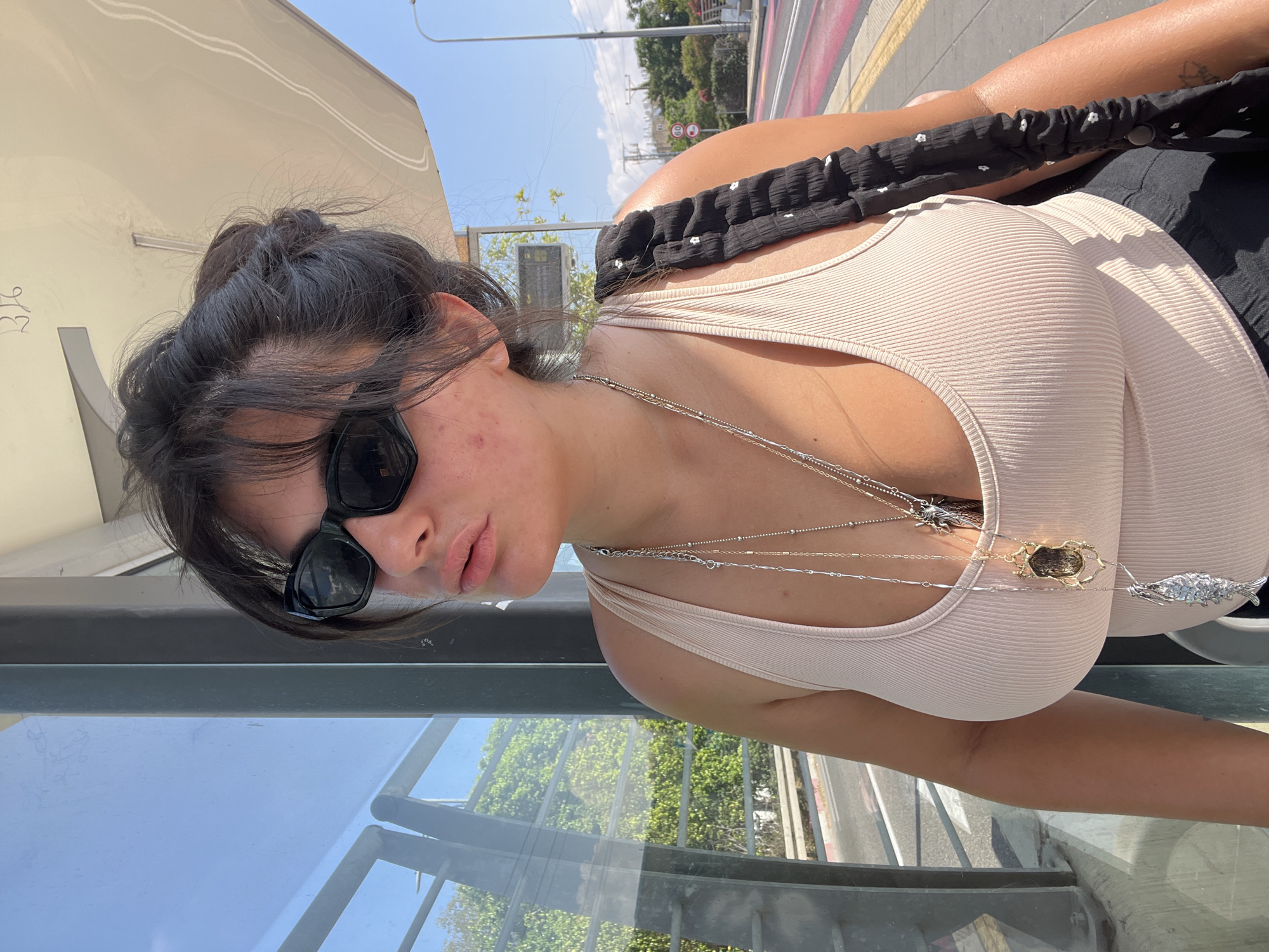 A woman wearing a beige tank top and sunglasses stands at a bus stop in Kiryat Motzkin, Israel. She has her dark hair pulled up in a messy bun and wears several necklaces with pendants. The bus stop features a glass enclosure and a beige awning. Behind her, there is a street with traffic signals and foliage. The weather appears sunny and clear.