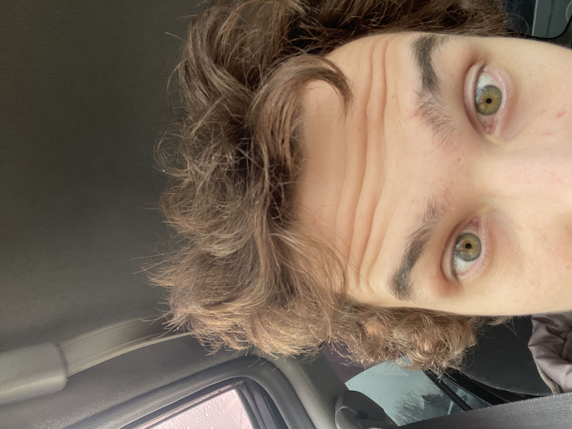 A selfie photograph taken from a close and high angle depicts a person with brown hair, green eyes, and forehead wrinkles. The setting appears to be inside a vehicle, evidenced by a car ceiling and a window with raindrops. There is no contextual information available to determine if the location is in Krasnoyarsk, Russia, or any other city. The image primarily features the individual's face and part of their upper body. No actions, interactions, or other people are visible.