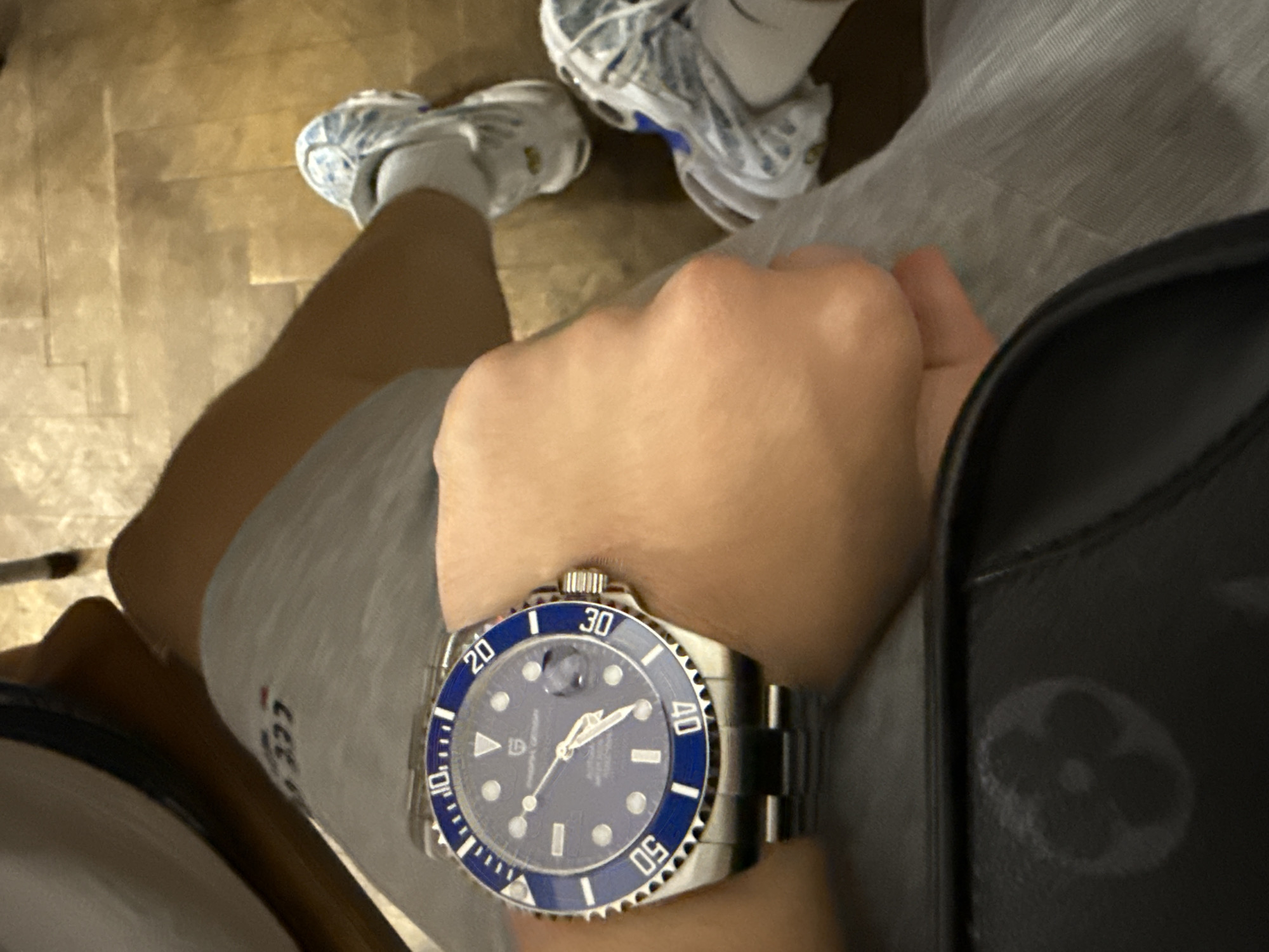 A high-angle, first-person perspective photograph captures a person's upper legs, feet, and a left arm wearing a wristwatch, against a background of a wooden floor. The individual appears to be seated or resting. They are wearing grey shorts and white socks, with a faint "333" mark visible on one sock. On their feet are white and blue sneakers, possibly Nike Air Max TNs, featuring visible air bubbles in the soles.
The prominent subject is a stainless steel wristwatch worn on the left wrist. It features a bright blue, serrated bezel with white minute markings and numbers (20, 30, 40, 50), and a matching blue dial. The dial displays luminous circular and rectangular hour markers, luminous hands, and a date window with a cyclops magnifier at the 3 o'clock position. Clearly visible text on the watch dial reads "PAGANI DESIGN," "AUTOMATIC," and "100M WATER RESISTANT." The time indicated by the watch hands is approximately 10:10.
To the right of the frame, a black bag, likely a shoulder or crossbody bag, is partially visible. It features a subtle, debossed or printed Louis Vuitton quatrefoil flower monogram pattern. The floor beneath the person is made of light brown wooden planks, suggesting an indoor setting. The overall scene depicts a casual moment, potentially in Burgas, Bulgaria, although the image itself lacks specific location identifiers for the city.
