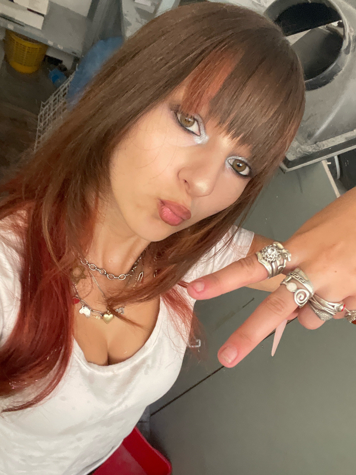 The image shows a young woman with long, reddish-brown hair and bangs. She is wearing a white t-shirt, a silver chain necklace with charms, and several silver rings on her fingers. She has makeup on, including prominent silver eyeshadow. The woman is posing with her hand extended, fingers pointed. The background is blurred and appears to be an indoor environment with some industrial or utility elements visible, such as a metal vent and shelving with containers. There is no indication of Raanana, Israel in the image.