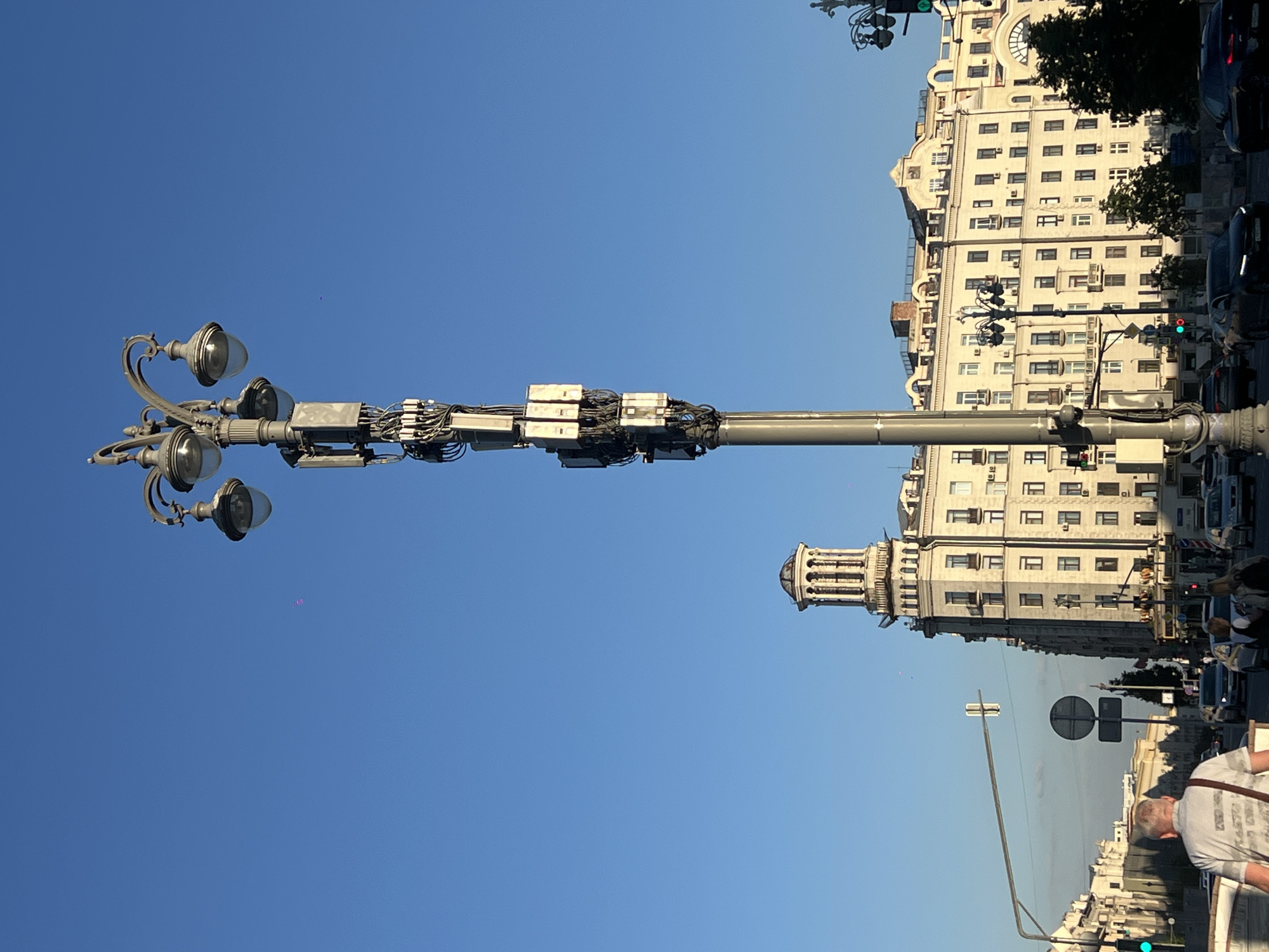 This image depicts an urban street scene in Moscow, Russia, specifically focusing on the area around Triumfalnaya Square and the Peking Hotel.
In the foreground, a tall, ornate grey lamppost dominates the center-right. It features four classic streetlights at its apex, each with a clear, globe-shaped diffuser. Below the lights, the pole is heavily populated with numerous rectangular electronic modules and a dense network of black cables, indicative of telecommunications or smart city infrastructure.
The sky above is clear and bright blue, occupying the upper half of the frame. A faint, small magenta speck is visible in the upper left quadrant.
In the midground, the prominent **Peking Hotel** (Гостиница "Пекин") at Bolshaya Sadovaya Ulitsa 5 is visible on the left. It is a large, light-colored, multi-story building characterized by a distinct classical tower with columns and a domed roof. The building's facade, illuminated by direct sunlight, shows numerous windows and balconies. To the right of the Peking Hotel, another large, multi-story building with a light-colored facade, numerous windows, and some roofline architectural details is visible.
Below the buildings, a multi-lane asphalt road extends across the scene. Several vehicles, including sedans of various colors (black, silver), are present on the road, some stationary, others in motion. Green traffic lights are visible on overhead gantries on both the left and right sides of the road. A circular black and white regulatory traffic sign is mounted on a pole on the left side of the street.
In the immediate left foreground, the back and right profile of a man with short grey hair, wearing a light-colored patterned polo shirt and a dark shoulder bag, is visible as he appears to walk on a pedestrian pathway or bridge. Further back, on the street level, several other pedestrians are discernible, including two women closer to the right side of the road. A dark green tree is visible on the far right, partially obscuring the building behind it. The scene is brightly lit by natural sunlight, suggesting a clear day.