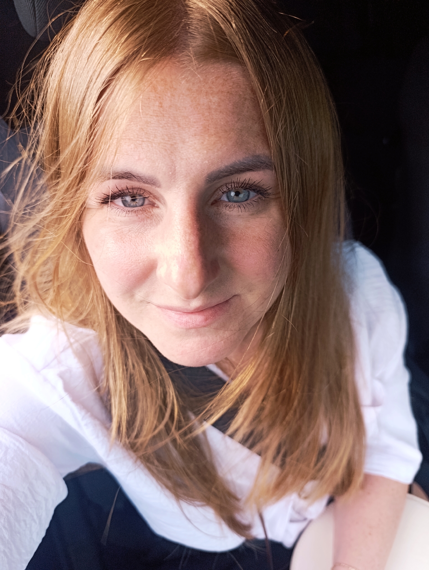 A young woman with blonde hair and blue eyes is pictured in a close-up, selfie-style shot. She is wearing a white collared shirt and appears to be sitting in a car or a similar enclosed space, with dark material visible in the background. Her face is turned towards the camera, and she has a slight smile. The lighting suggests an outdoor setting, possibly with sunlight casting shadows on her face.