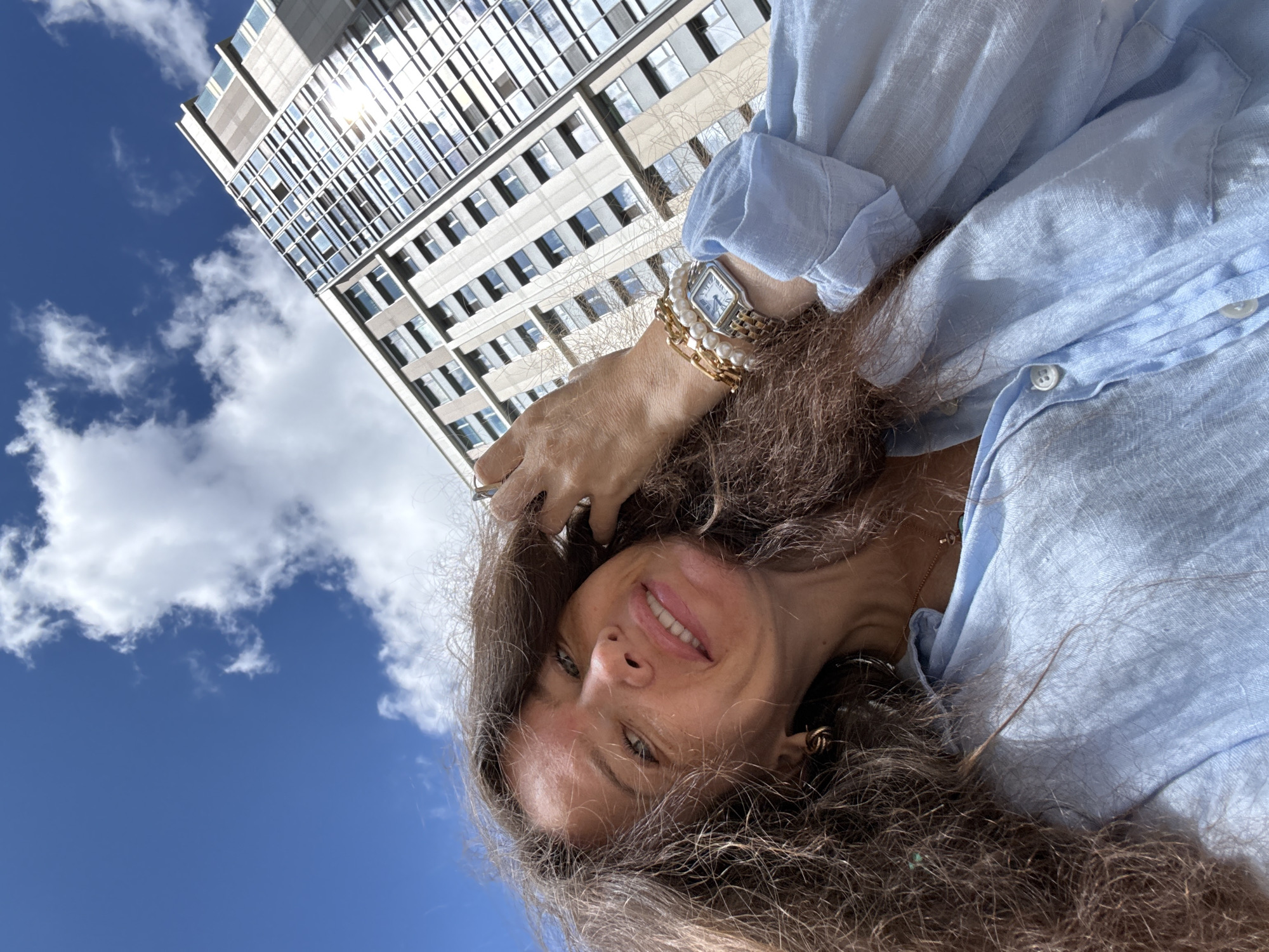 A low-angle, close-up, selfie-style photo shows a woman with long, curly brown hair and fair skin. She is wearing a light blue linen shirt and has a pearl and gold bracelet on her left wrist. She is smiling and touching her hair. Behind her, a modern, tall building with many windows rises against a bright blue sky with scattered white clouds. The building appears to be in an urban setting, possibly Moscow, Russia. The sunlight reflects off the windows of the building.