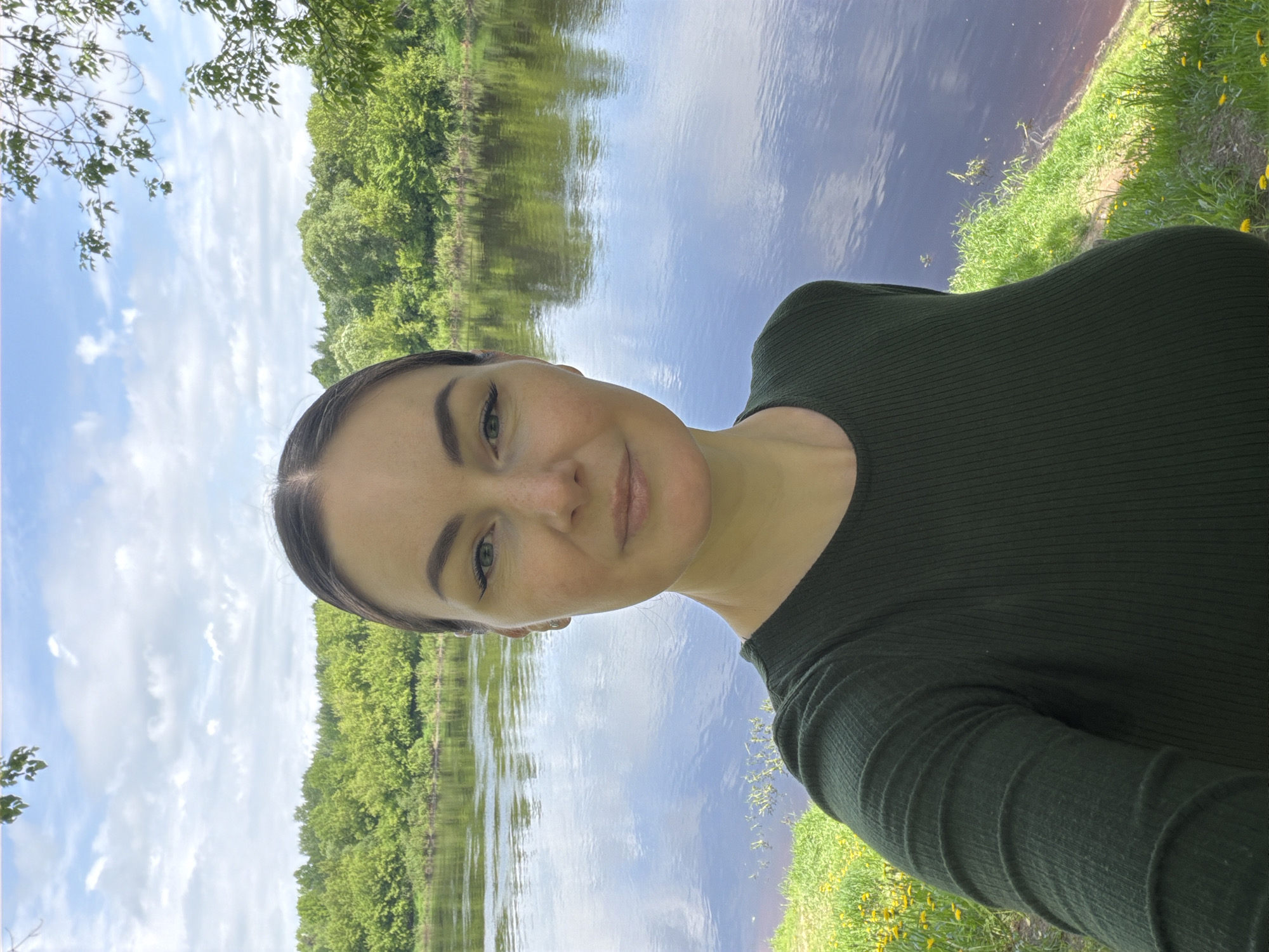The image captures a woman in Unknown, Unknown, posing for a selfie outdoors. She is wearing a dark green, long-sleeved shirt. The background features a body of water with reflections of trees and sky. The sky is blue with scattered clouds. Vegetation lines the edge of the water, including grass and yellow flowers.