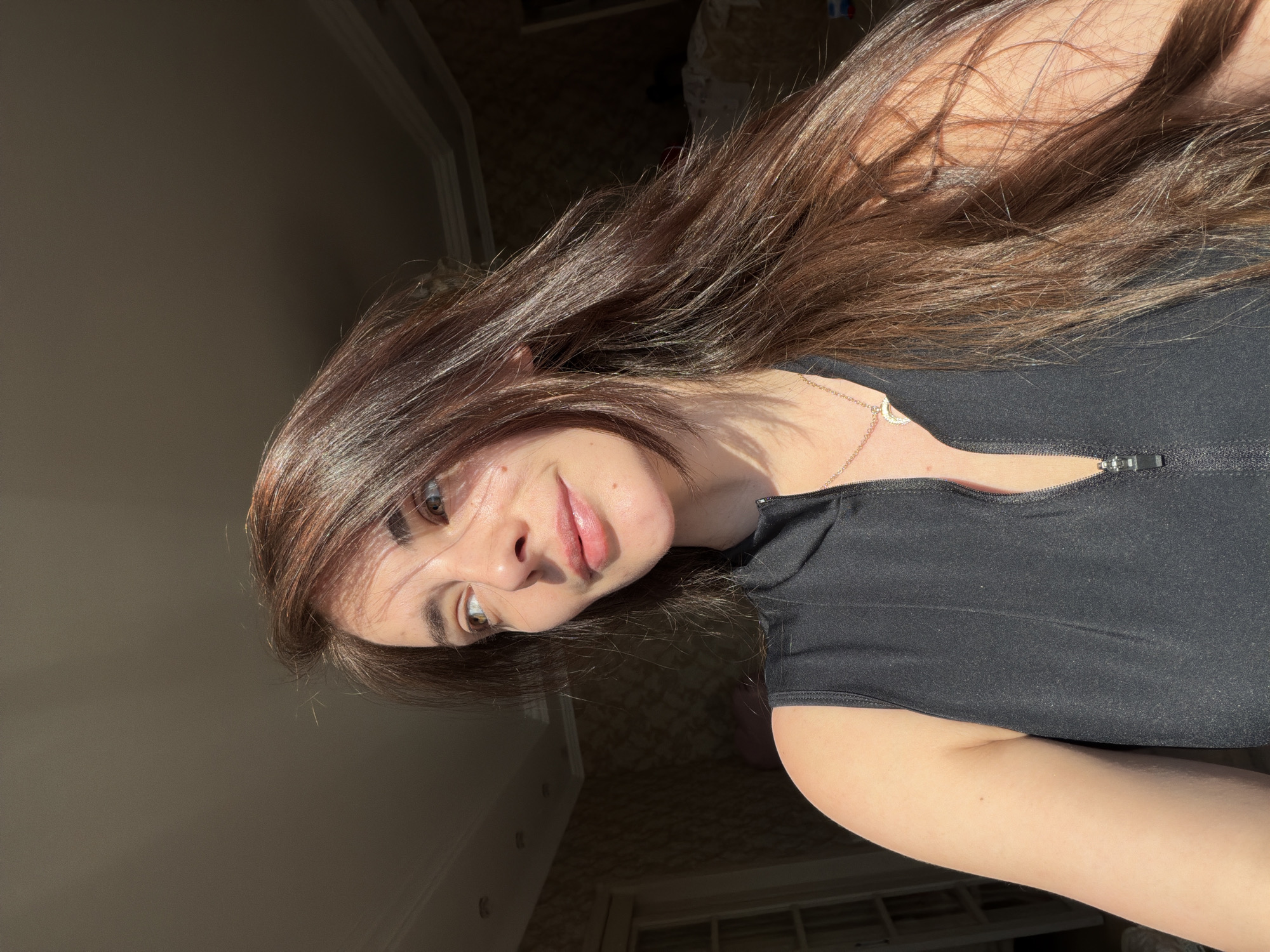 A woman with long, dark brown hair is pictured from the chest up, looking to her left. She is wearing a black, sleeveless top with a zipper down the front. Sunlight streams in from the left, illuminating her face and hair. The background appears to be an indoor setting with patterned wallpaper and molding on the ceiling.