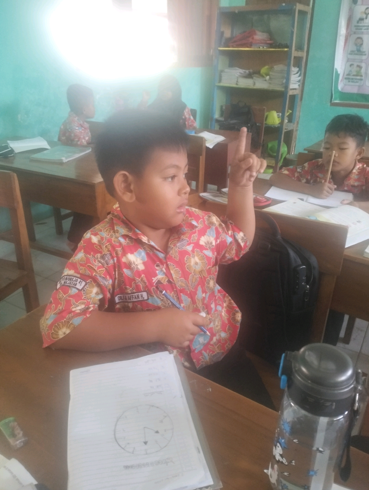 In a classroom in Blabakan, Indonesia, a young boy wearing a red and white patterned shirt with blue accents raises his right index finger while looking to his right. He is seated at a wooden desk, in front of him is an open notebook with a circular diagram, and a pen. A black backpack is visible behind him. To his right, another young boy in a similar shirt is visible, leaning over a notebook and holding a thin stick. The classroom is brightly lit by natural light from a window, casting a glow on the pale blue wall. Bookshelves filled with papers and books are visible in the background, along with wooden chairs and tables. A clear water bottle with a black lid is also on the desk in the foreground.