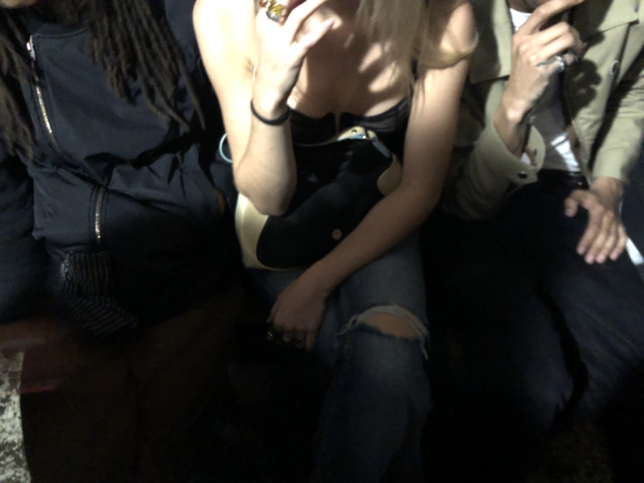 Three people are closely seated. The person on the left has dark dreadlocks and wears a black jacket. The individual in the middle has blonde hair, a black top, jeans with a rip at the knee, and a black and beige bag across their lap. They wear rings and a black bracelet. The person on the right wears a light-colored jacket and dark pants. All three appear to be in a dimly lit indoor setting.