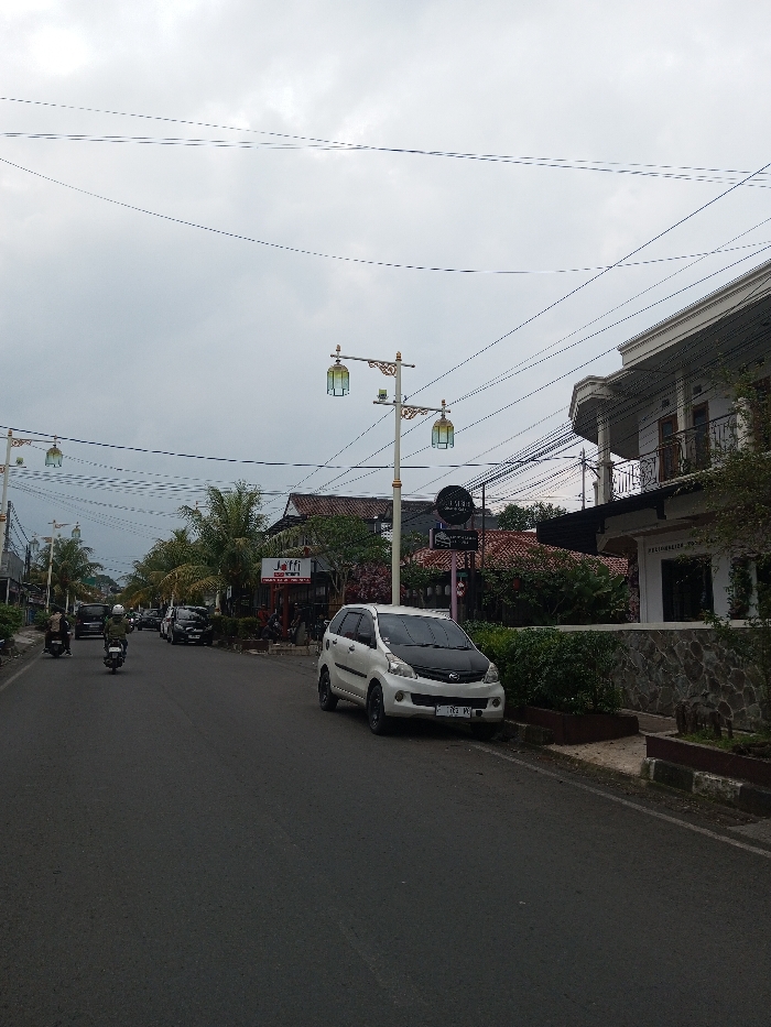 A street view features a white car parked near a sidewalk. On the left, motorcycles move along the road. Buildings and various signs are visible along the street, including "Jaffi". Decorated street lights line the road under a cloudy sky filled with numerous electrical wires. Some greenery is planted along the street's edge.