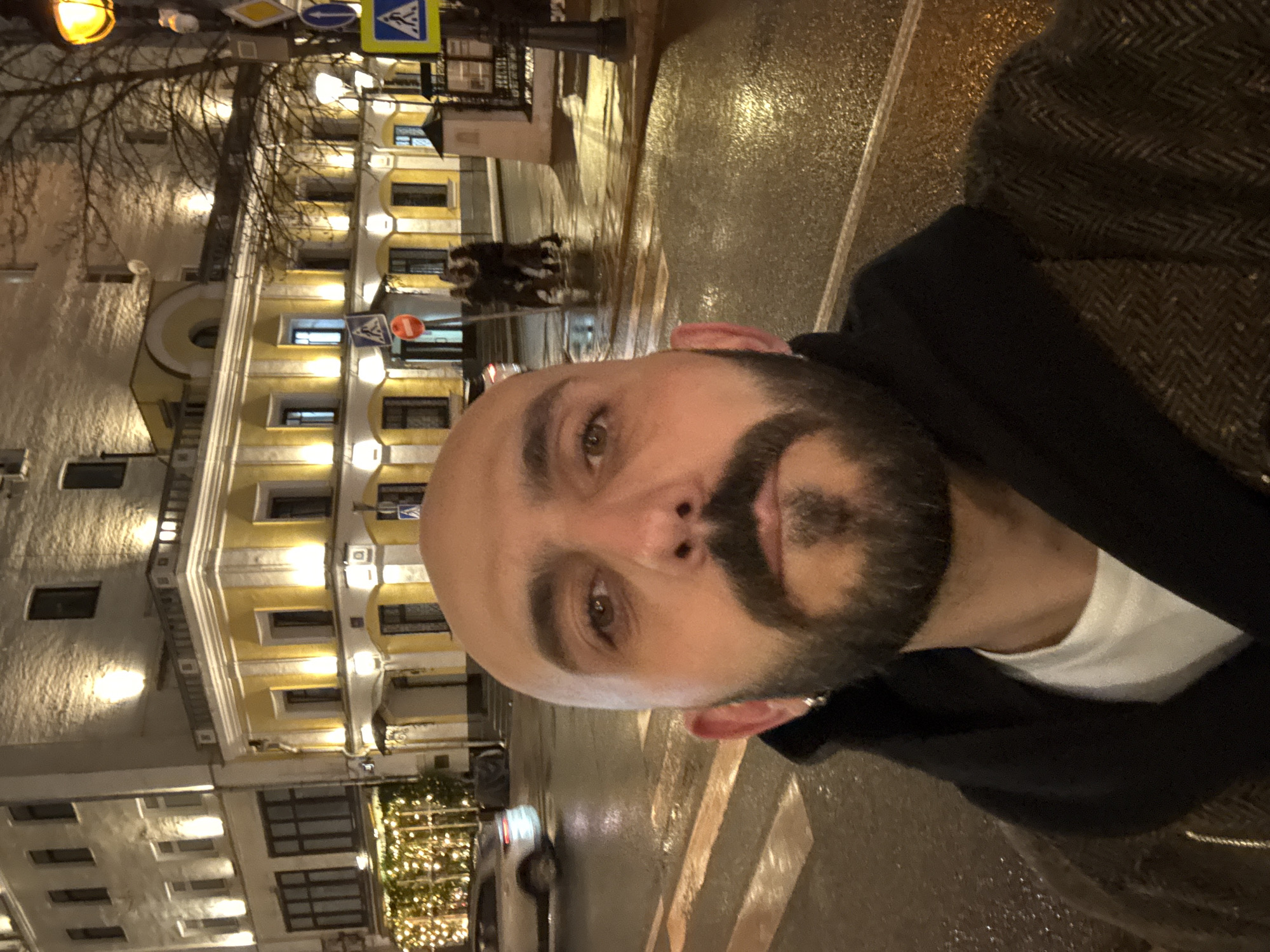 A selfie captured at night in Moscow, Russia, shows a bald, bearded man in a white shirt and dark jacket standing on a wet, striped crosswalk. Behind him, an illuminated yellow building contrasts with bare trees. Traffic signs are visible above him. Christmas lights decorate a tree in the lower left. A car passes in the background. The man's face is in sharp focus.