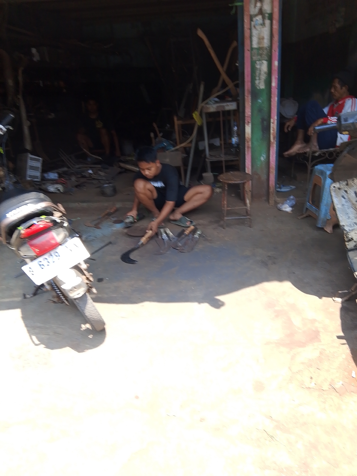 In Gedangan, Indonesia, a young person is shown working in a dimly lit open-air workshop. The individual, wearing a black t-shirt and shorts, is crouched on the ground, sharpening a large curved blade, possibly a machete or sickle, with a grinding tool. The workshop appears cluttered with various tools, metal scraps, and a motorcycle parked to the left of the frame. The motorcycle has a white license plate with the number "B 6339 VV". In the background, another person is seated, wearing a red and white striped shirt and dark pants, appearing to be resting or observing. The ground is concrete, and the overall atmosphere suggests a functional, possibly traditional, metalworking or repair shop. Sunlight casts strong shadows across the ground, indicating it is daytime.