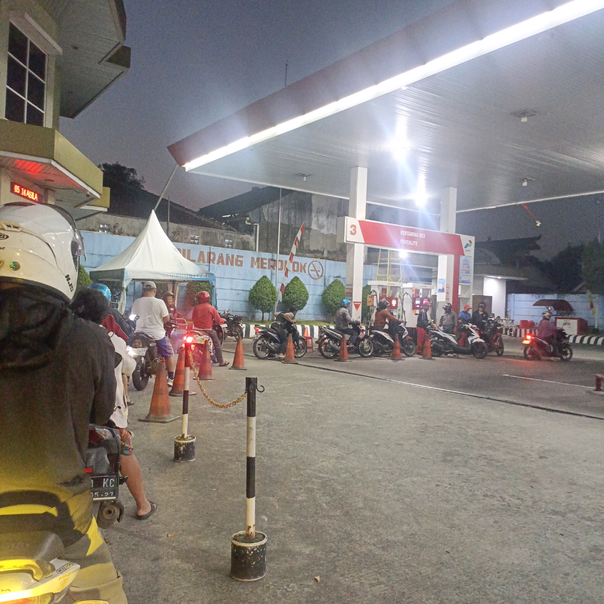 This image depicts a scene at a Pertamina gas station in Salatiga, Indonesia, during twilight. The station features a covered forecourt illuminated by long fluorescent lights. A queue of motorcycles and their riders is visible, waiting for fuel.
In the foreground, the back of a person wearing a black hoodie and a white helmet is prominent, seated on a yellow-accented motorcycle. To their right, another person in a white t-shirt and blue shorts is seated on a motorcycle. Several other individuals on motorcycles are spread across the forecourt, some appearing to be in line for service.
Behind the queue, a white tent is set up, and a blue wall with a sign that reads "DILARANG MEROKOK" (No Smoking) is visible, along with stylized depictions of trees. The gas pumps are positioned under the covered area, with their electronic displays visible.
The ground is paved asphalt. Orange traffic cones and a chain barrier with black and white striped posts are present, delineating areas and guiding traffic. The overall atmosphere suggests a routine evening at a local gas station, with people engaged in the common activity of refueling their vehicles. The sky is a dusky blue, indicating the transition from day to night.