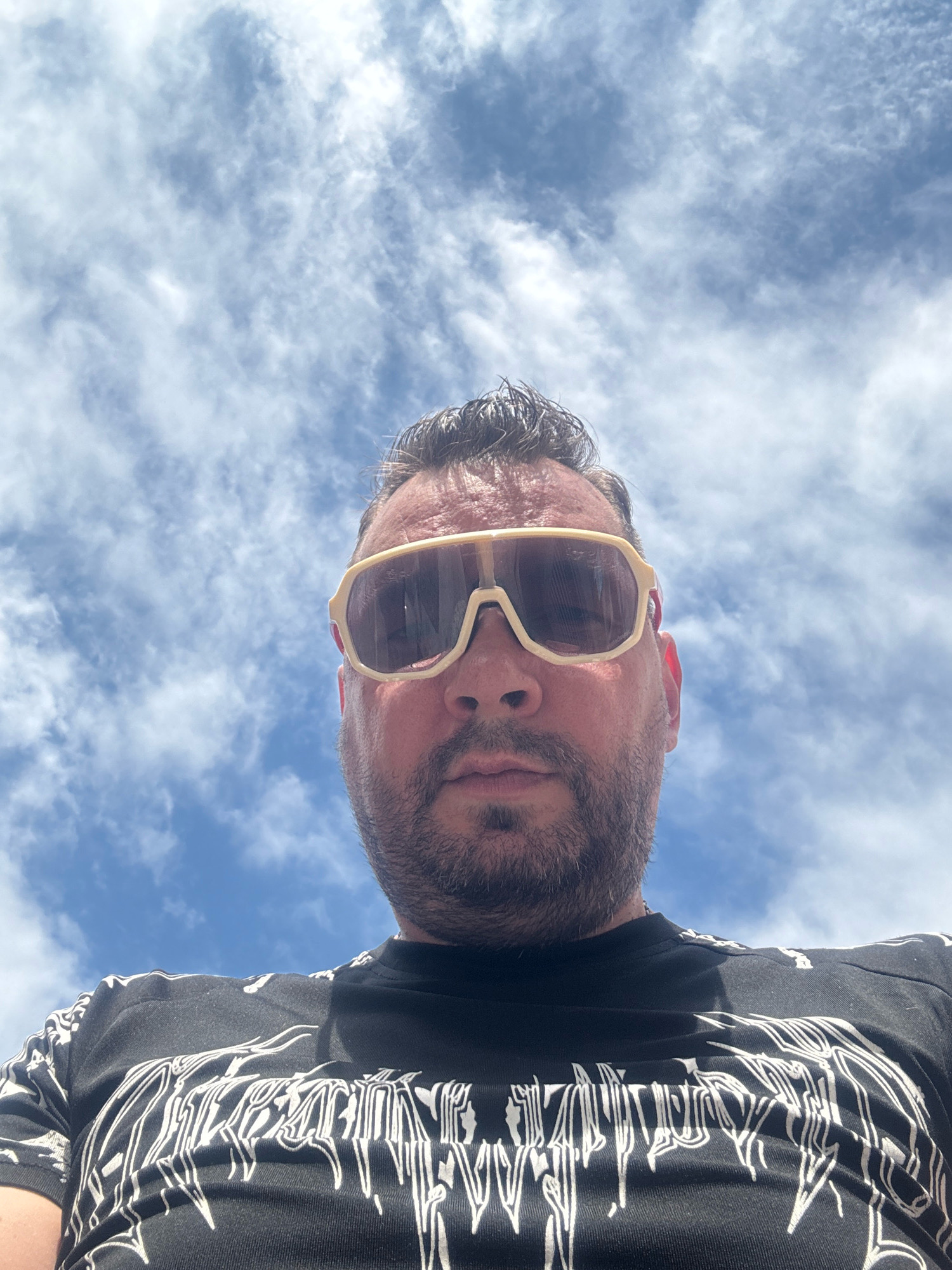 A man is captured from a low angle, looking up into a cloudy sky. He is wearing a black t-shirt with a white, gothic-style graphic print and large, cream-colored sunglasses with tinted lenses. His hair is dark and styled upwards, and he has a beard. The background is dominated by a bright blue sky with scattered white, wispy clouds, suggesting an outdoor setting, possibly during the daytime. There are no discernible landmarks or architectural features visible in this image to indicate a specific location within Nanhai District, China.