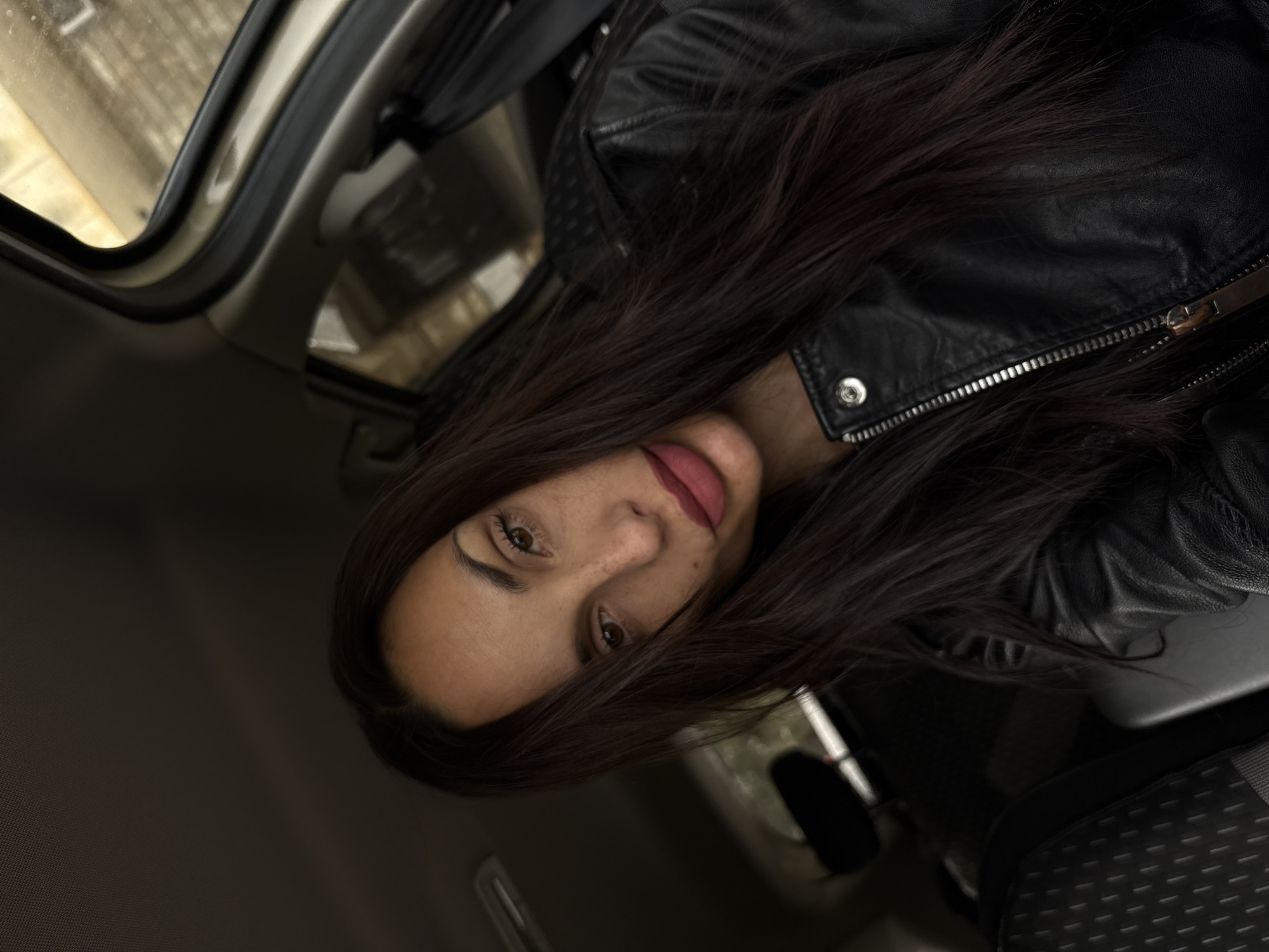 A woman with long, dark hair and brown eyes is sitting in the driver's seat of a car, wearing a black leather jacket. She is looking directly at the camera. The interior of the car is visible, with the car door frame on the left and the seat to the right. The environment outside the car is blurry and indistinct. The location is in the city of Pleven, Bulgaria.